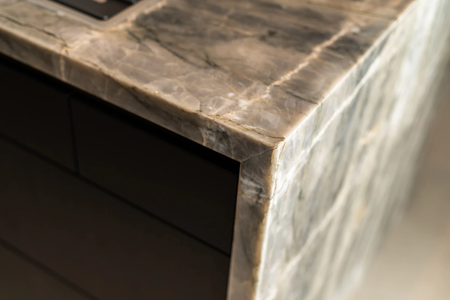 Close-up van de hoek van een kwartsiet werkblad, met een doorschijnend grijs-beige gemarmerd patroon en gladde randen. Onder het gepolijste stenen oppervlak zijn donkere kastladen zichtbaar.