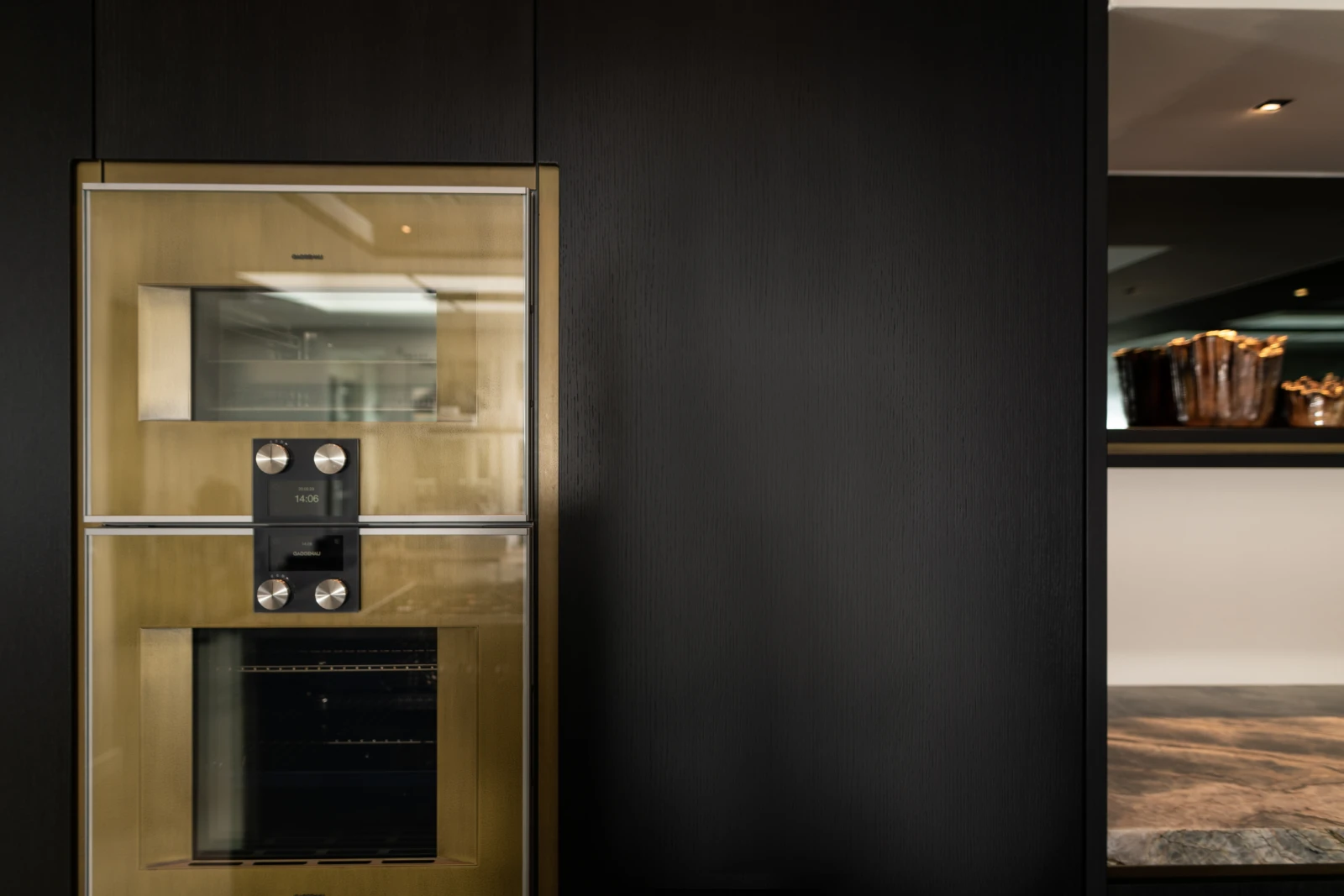 Een moderne keuken met schier kookeiland heeft een ingebouwde oven en magnetron met een geborsteld gouden afwerking in een donkere kast. Een strak kwartsiet werkblad en decoratieve items maken de stijlvolle look compleet.