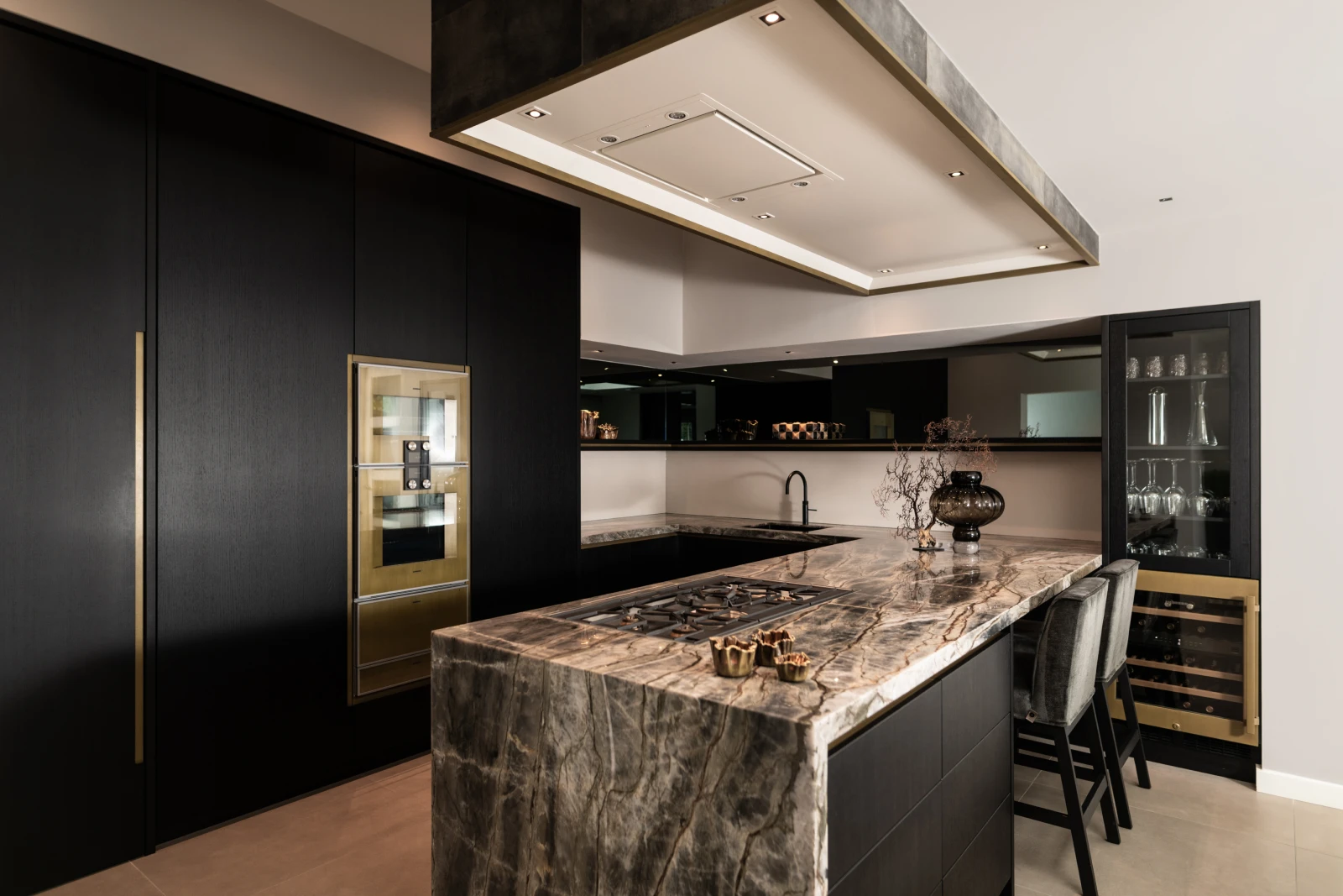 Moderne keuken met schier kookeiland, donkere kasten, kwartsiet werkblad, ingebouwde kookplaat en oven, twee barkrukken, glazen wijnkoeler en decoratieve vazen onder sfeervolle verlichting.