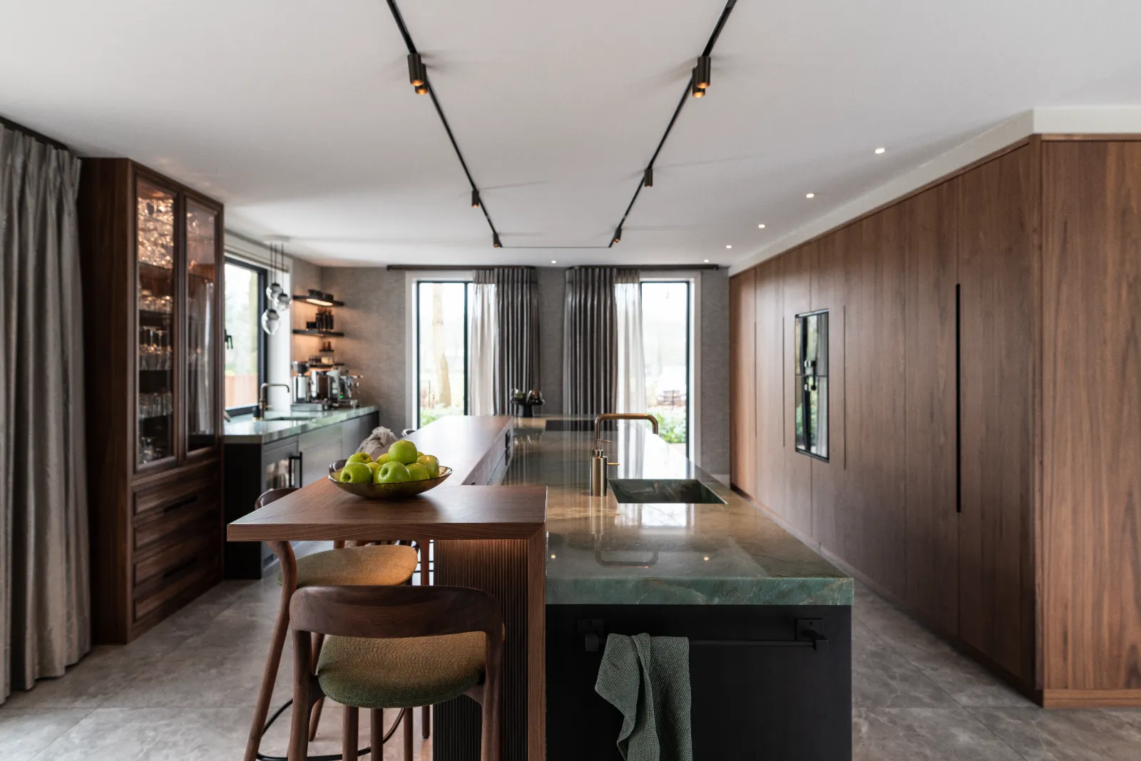 Moderne keuken met houten lambrisering, marmeren eiland, barkrukken en hanglampen. Grote ramen met gordijnen laten natuurlijk licht binnen op het aanrecht van deze exclusieve keuken, waar een schaal met groene appels voor een frisse toets zorgt.