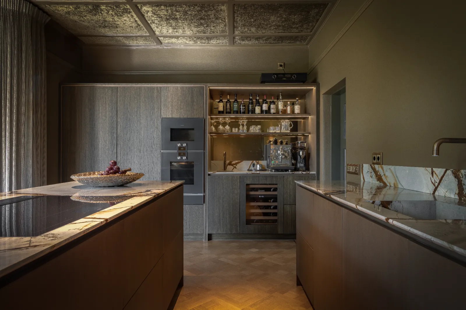 Moderne keuken met donkerhouten kasten, ingebouwde apparatuur en verlichte planken met glaswerk en flessen. Marmeren aanrechten en een schaal met druiven staan op een eiland en tonen de elegantie van exclusieve keukens.
