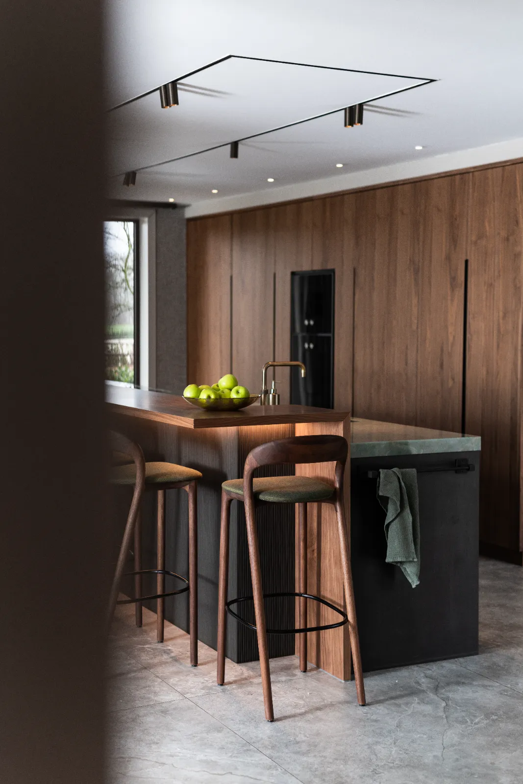 Moderne keuken met houten kastjes, een zwart eiland, twee houten barkrukken, een gootsteen en een bos groene appels op het aanrecht. Deze minimalistische ruimte met daglicht en een handdoek op de vaatwasser weerspiegelt de elegantie van exclusieve keukens.