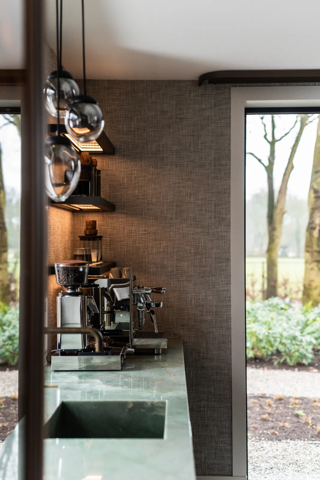 Modern aanrecht met een groen oppervlak, espressomachine en koffiemolen. Hanglampen hangen boven deze exclusieve keukensetting, planken tonen boeken en decor, en grote ramen onthullen een weelderige tuin met bomen.