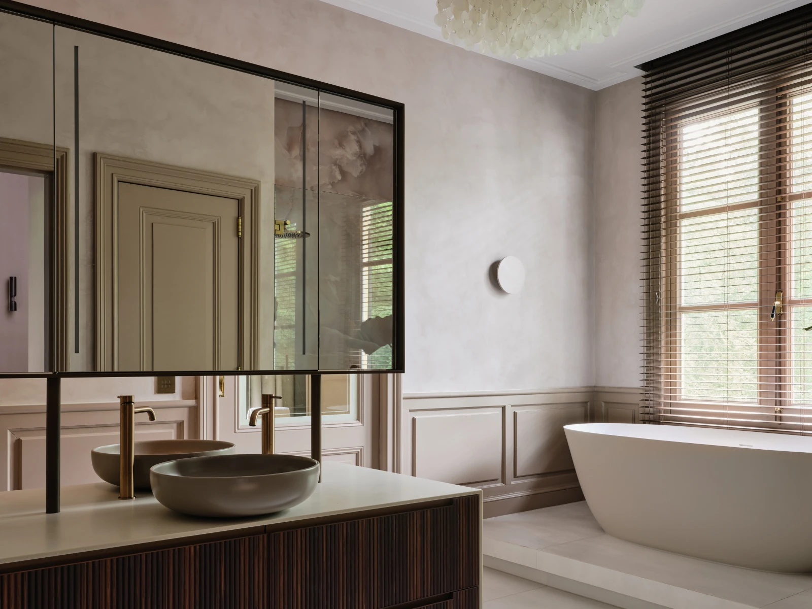 Een moderne badkamer in de stijl van een interieur herenhuis, met een grote spiegel, dubbele wastafels, gouden kranen, een vrijstaand wit bad, hoge ramen met luxaflex, taupe muren en zacht natuurlijk licht.