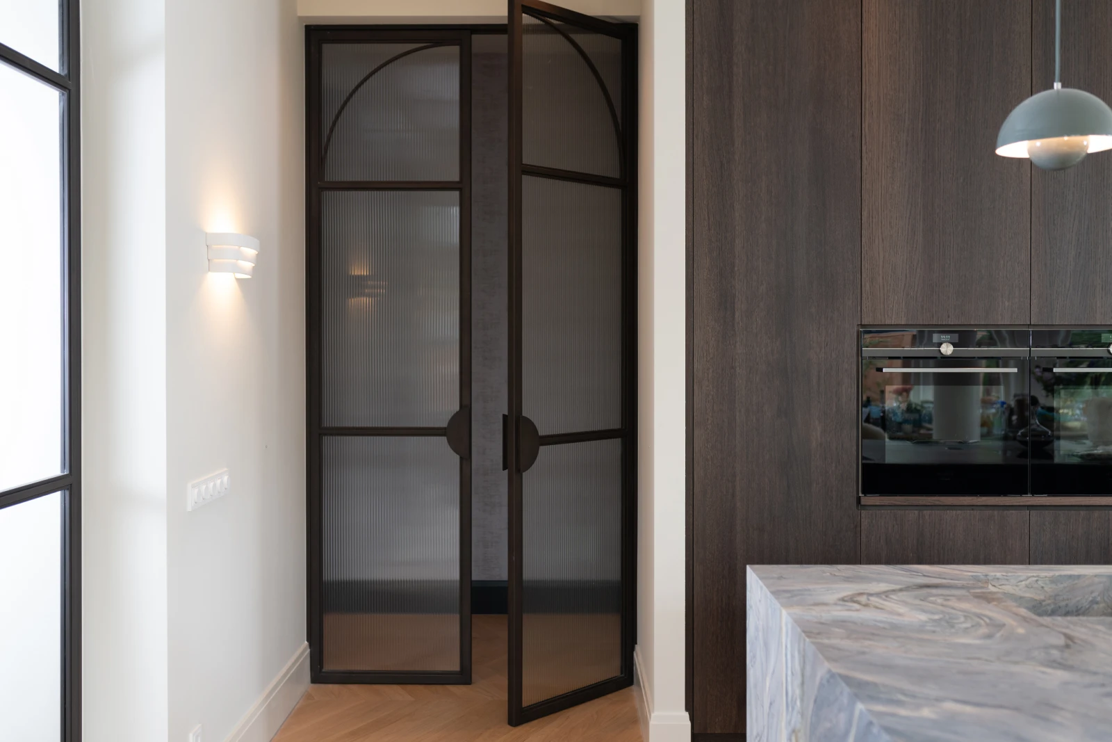 Een moderne keuken in een monumentaal pand heeft een zwarte glazen dubbele deur in een witte muur met een verlichtingsarmatuur. Daarnaast is in een donkerhouten wand een inbouwoven geplaatst en is een deel van een marmeren aanrecht zichtbaar.