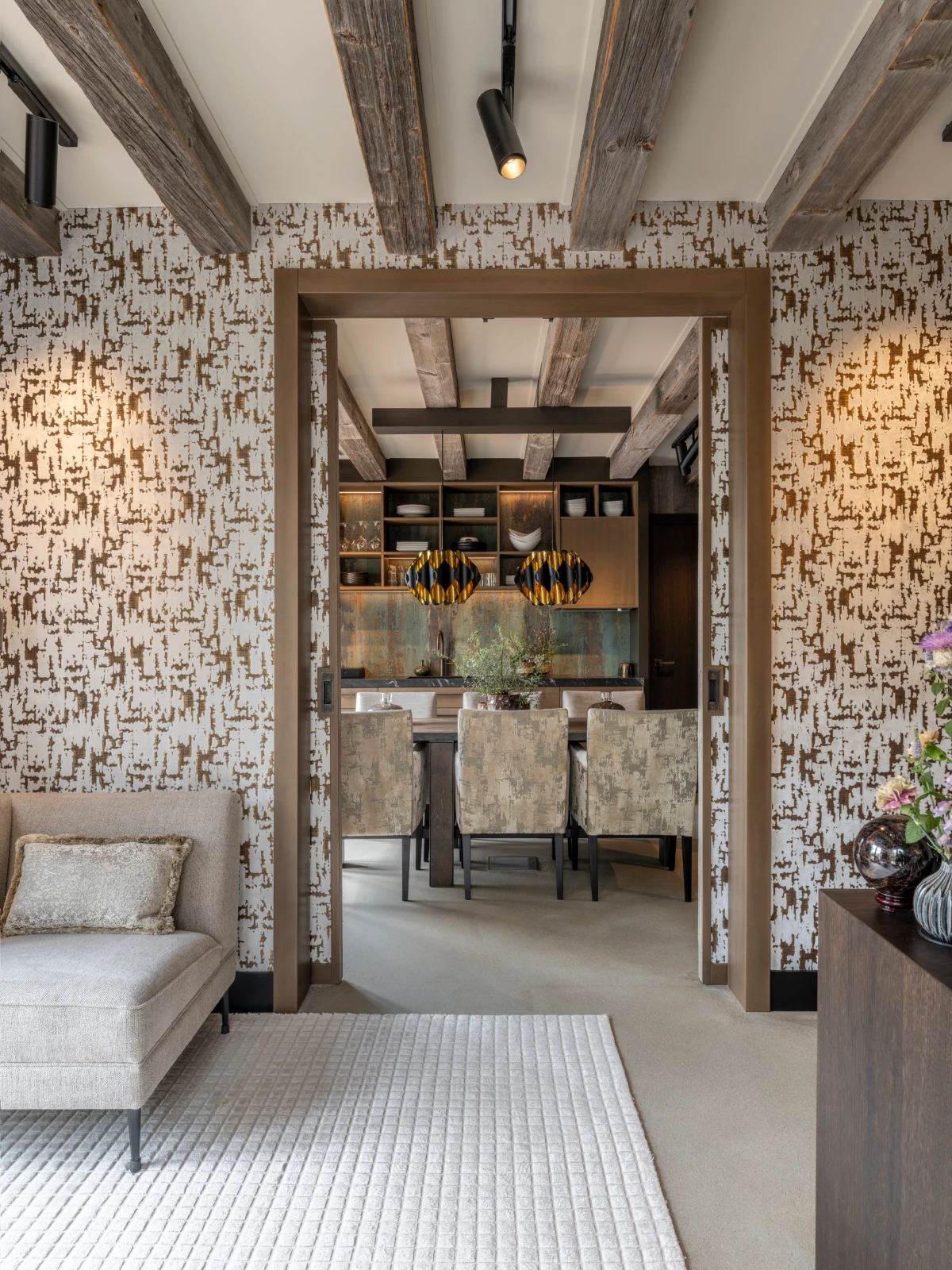 Een moderne villa interieur heeft een woonkamer met patroonbehang, houten balken en een lichte bank die overloopt in een eethoek met een grote tafel, gestoffeerde stoelen, zwarte hanglampen en een met bloemen gevulde vaas op de bijzettafel.