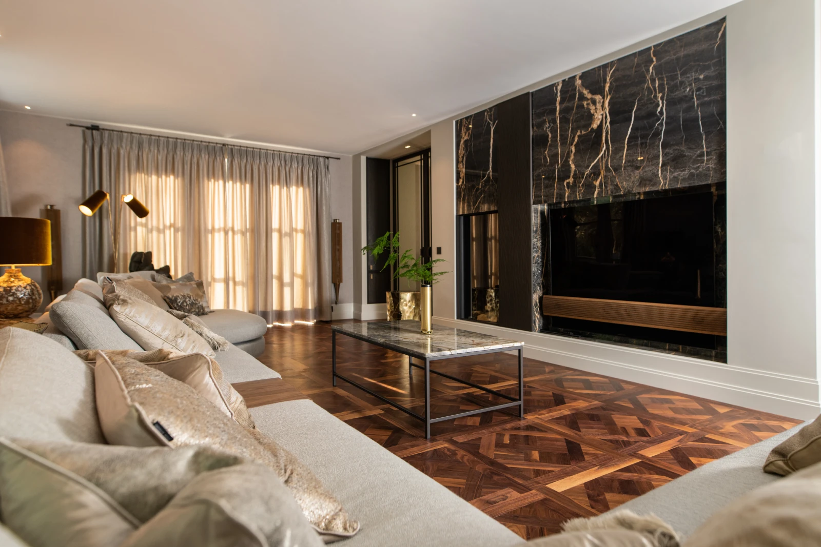 Luxe maatwerk interieur met een moderne woonkameropstelling: een royale hoekbank, geometrisch houten vloerpatroon en een op maat ontworpen marmeren wand met geïntegreerde haard en tv.