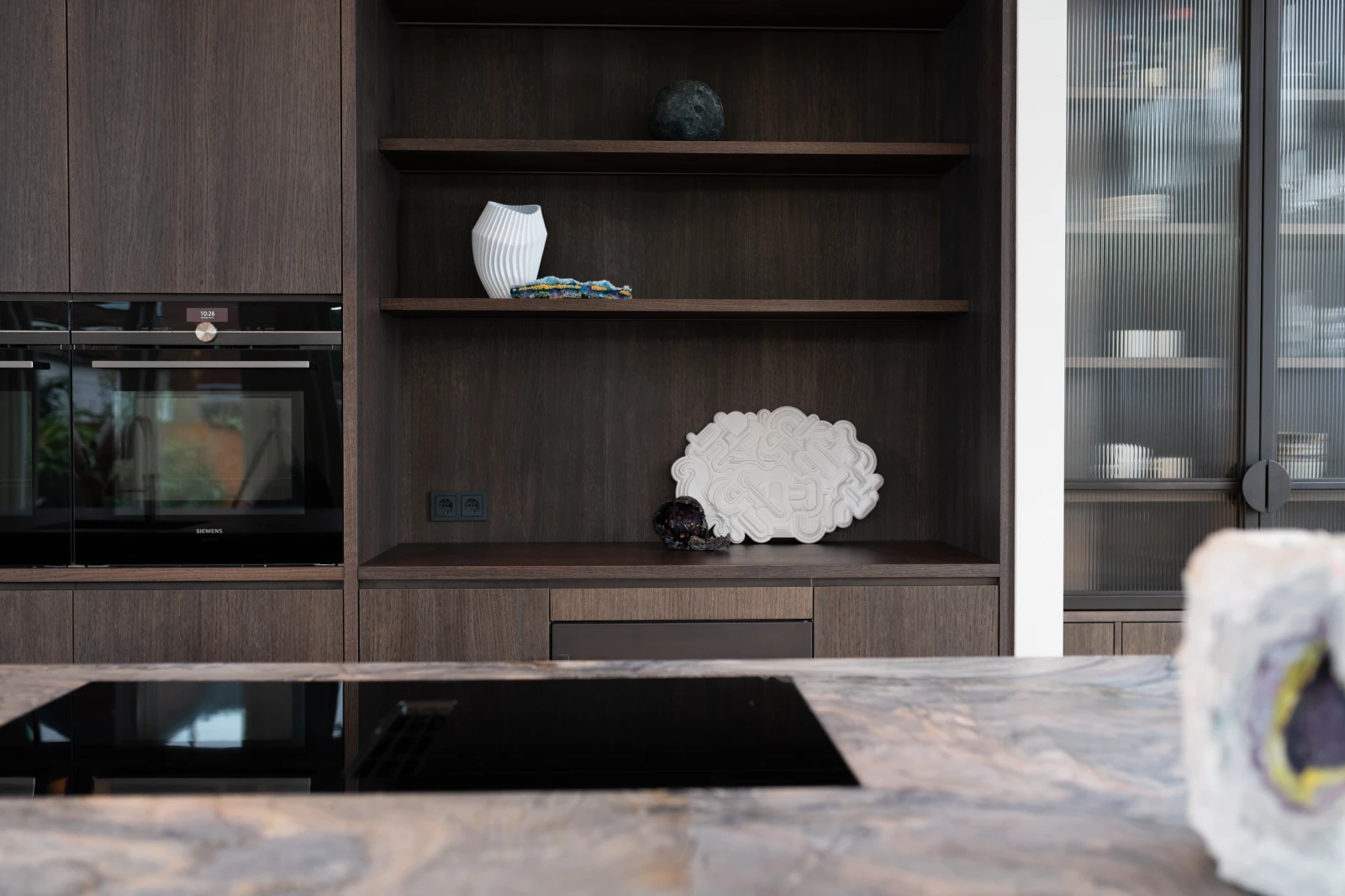 Een moderne eiken keuken met donkerhouten kasten, open planken met een witte vaas en decoratieve voorwerpen, en een gemarmerd kwartsiet keukenblad met een zwarte kookplaat op de voorgrond.