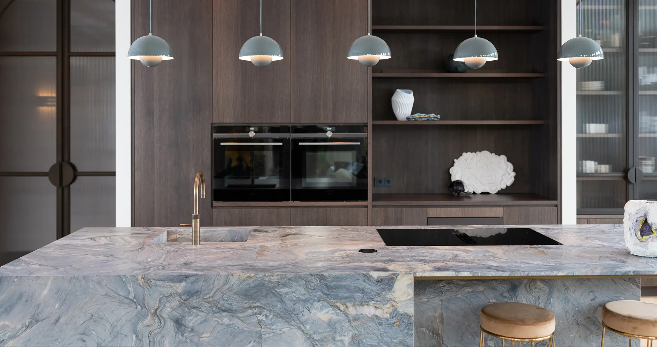 Moderne eiken keuken met een kwartsiet keukenblad, ingebouwde oven, minimalistische houten kasten, open planken met decor en vier hanglampen boven het marmeren eiland. Bij het eiland staat een gouden kruk.