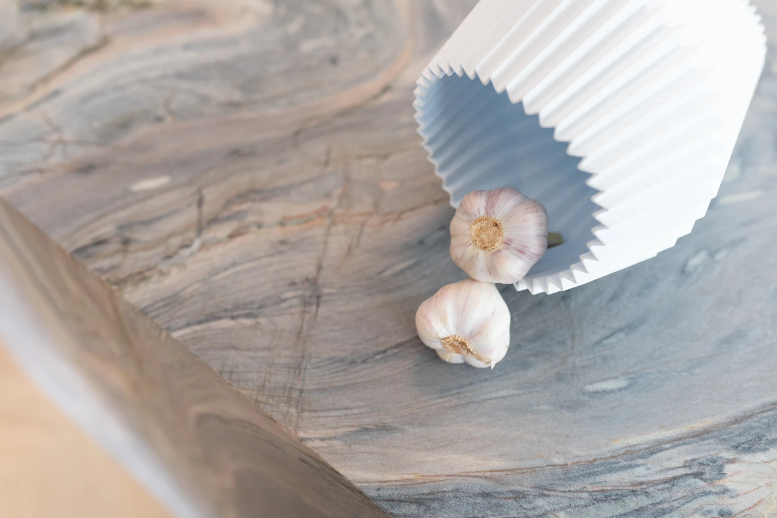 Twee knoflookbollen komen gedeeltelijk uit een witte, geribbelde bak op een marmeren kwartsiet keukenblad met elegante grijze en beige krullen.