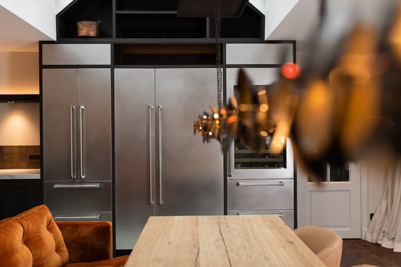 Een moderne industriele keuken met grote roestvrijstalen koelkasten, een eiken fineer keuken eettafel en een zacht oranje stoel op de voorgrond. Vage hanglampen rechtsvoor creëren een gezellige sfeer.