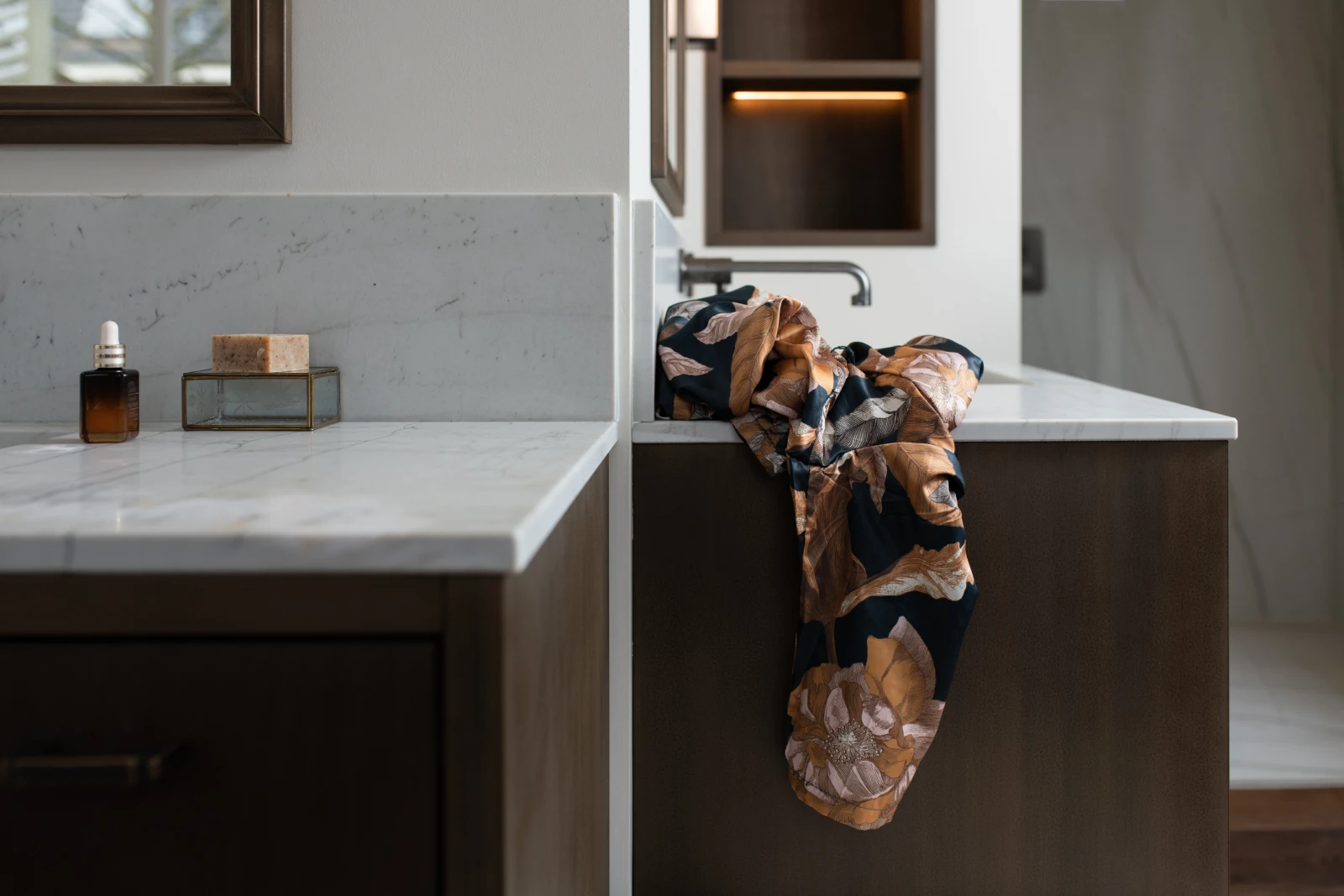 Een gebloemde badjas is gedrapeerd over de rand van een marmeren aanrecht. Vlakbij staan een bruin flesje met een druppelaar en een glazen bakje met een stuk zeep op een ander marmeren aanrecht.
