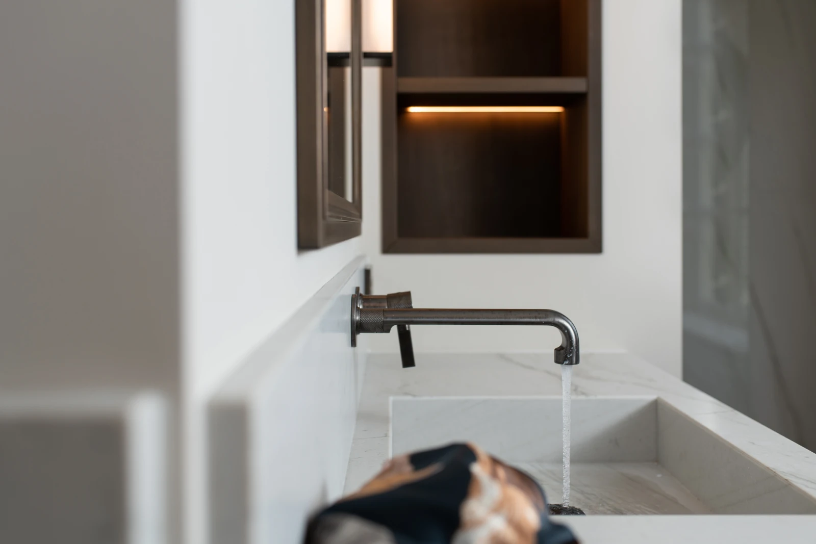Een moderne badkamer met een aan de muur gemonteerde kraan die water in een rechthoekige witte marmeren wastafel giet, met planchetten en een spiegel aan de muur op de achtergrond.
