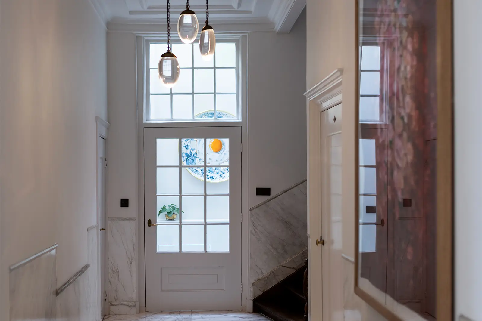 Een lichte hal met marmeren muren, een glazen deur, drie hangende hanglampen en een raam met blauw en oranje decoratief glas. Een kleine plant staat op een plank bij het raam.