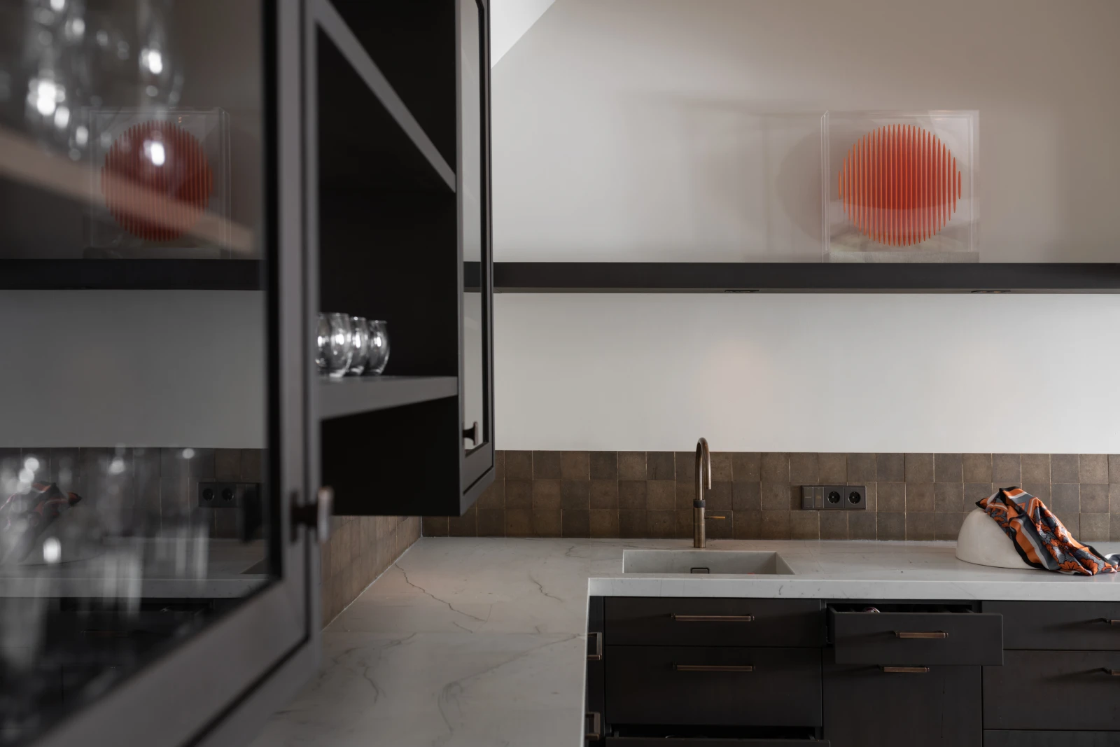 Een moderne, industriële keuken met donkere kasten en een marmeren aanrechtblad is voorzien van een gootsteen en kraan. Glazen staan op de plank, terwijl een decoratieve rode bol in een doorzichtige doos de ruimte boven het aanrecht accentueert.