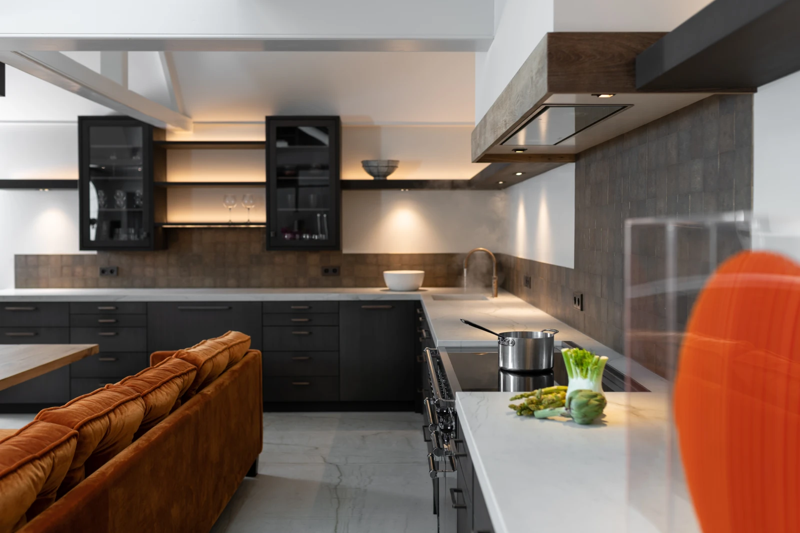 Moderne industriële keuken met donkere kasten, witte werkbladen en verlichting onder de kasten. Een Falcon Mercury 1200 staat op het fornuis, een pan staat te pruttelen en verse groenten liggen op het aanrecht. Een oranje sofa is gedeeltelijk zichtbaar op de voorgrond.