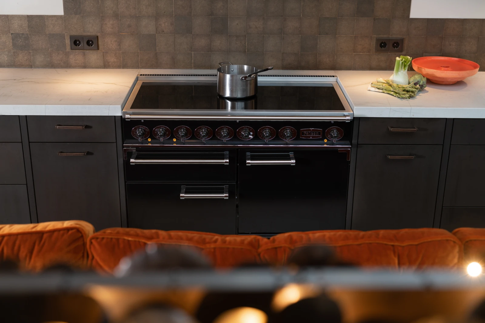 Een moderne industriele keuken met een zwart Falcon Mercury fornuis en oven, een pan op het fornuis en selderij en een oranje kom op het witte aanrecht. Donkere kasten, eiken fineer accenten en een betegelde achterwand maken het plaatje compleet.