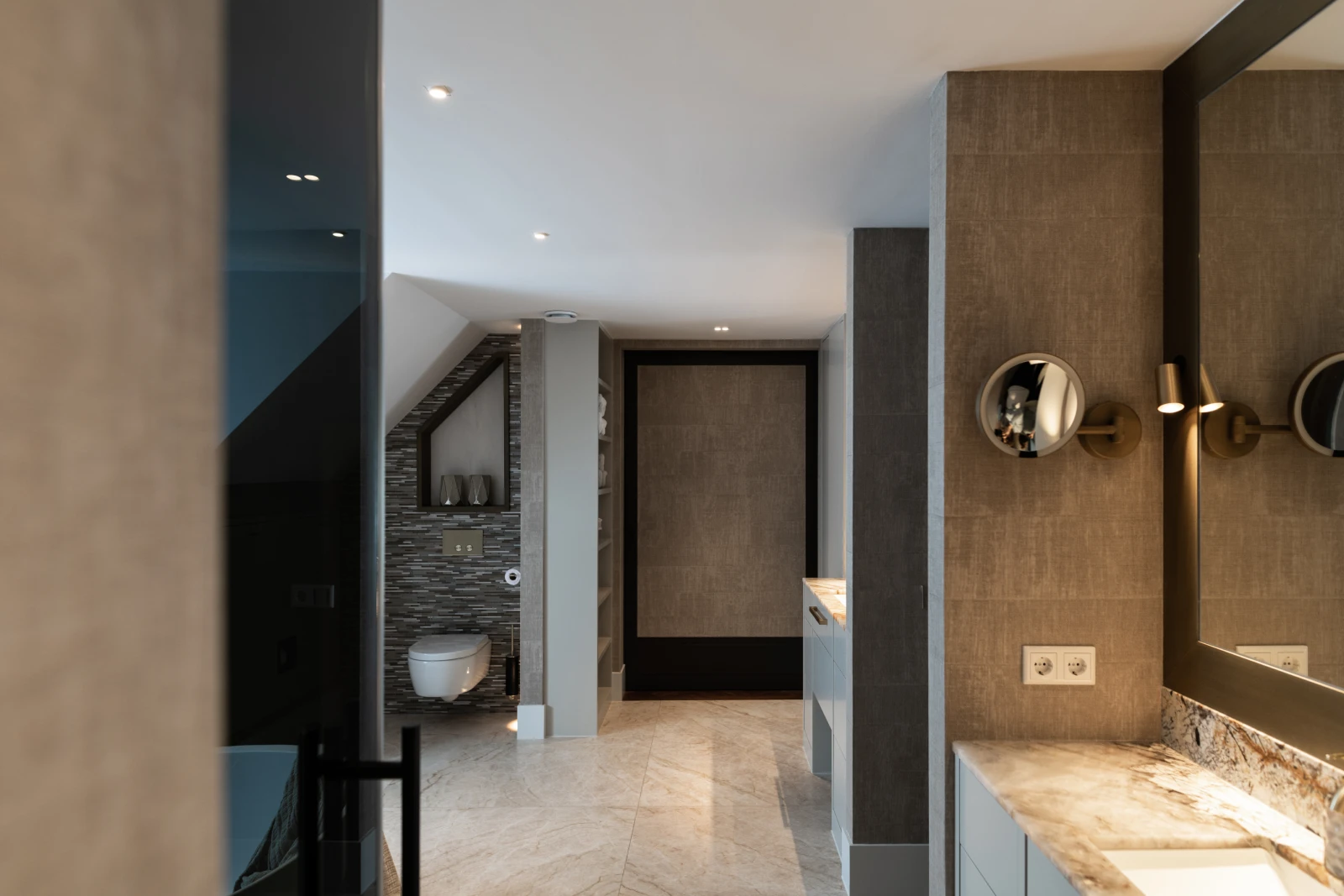 Moderne badkamer met beige tegelvloeren, grijze muren, een wandtoilet in een verzonken gedeelte, dubbele wastafels met marmeren werkbladen en ronde wandlampen naast een grote spiegel. De ruimte ziet er strak en eigentijds uit.