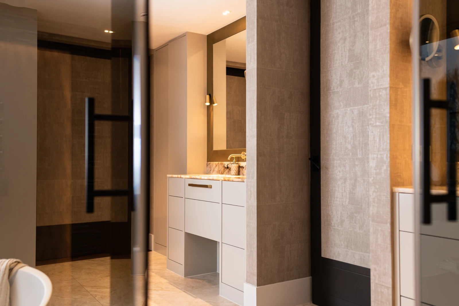 Moderne badkamer met beige gestructureerde muren, een grote spiegel boven een lichtgekleurd badmeubel met wastafel, marmeren aanrechtblad, zachte verlichting en strakke, minimalistische designelementen.