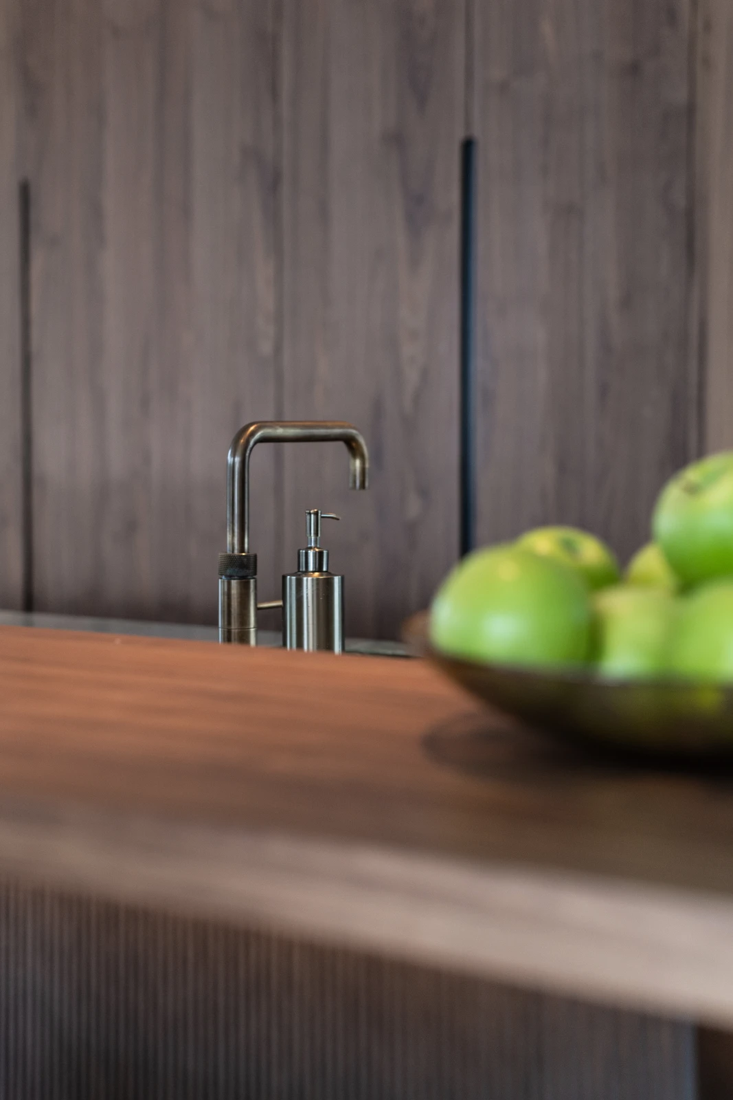 Een modern aanrecht met een metalen kraan en zeepdispenser, met een vage schaal met groene appels op de voorgrond en donkere houten kasten op de achtergrond.