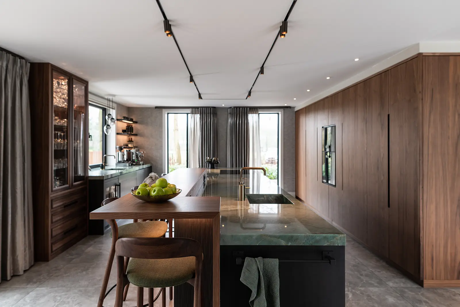 Moderne keuken met houten lambrisering, een opvallend groen kwartsiet keukenblad eiland, barhoog zitgedeelte met twee krukken, hangende tracklights en een schaal met groene appels. Grote ramen en planken bieden natuurlijk licht en opbergruimte. De Gaggenau kookplaat maakt de ruimte compleet.