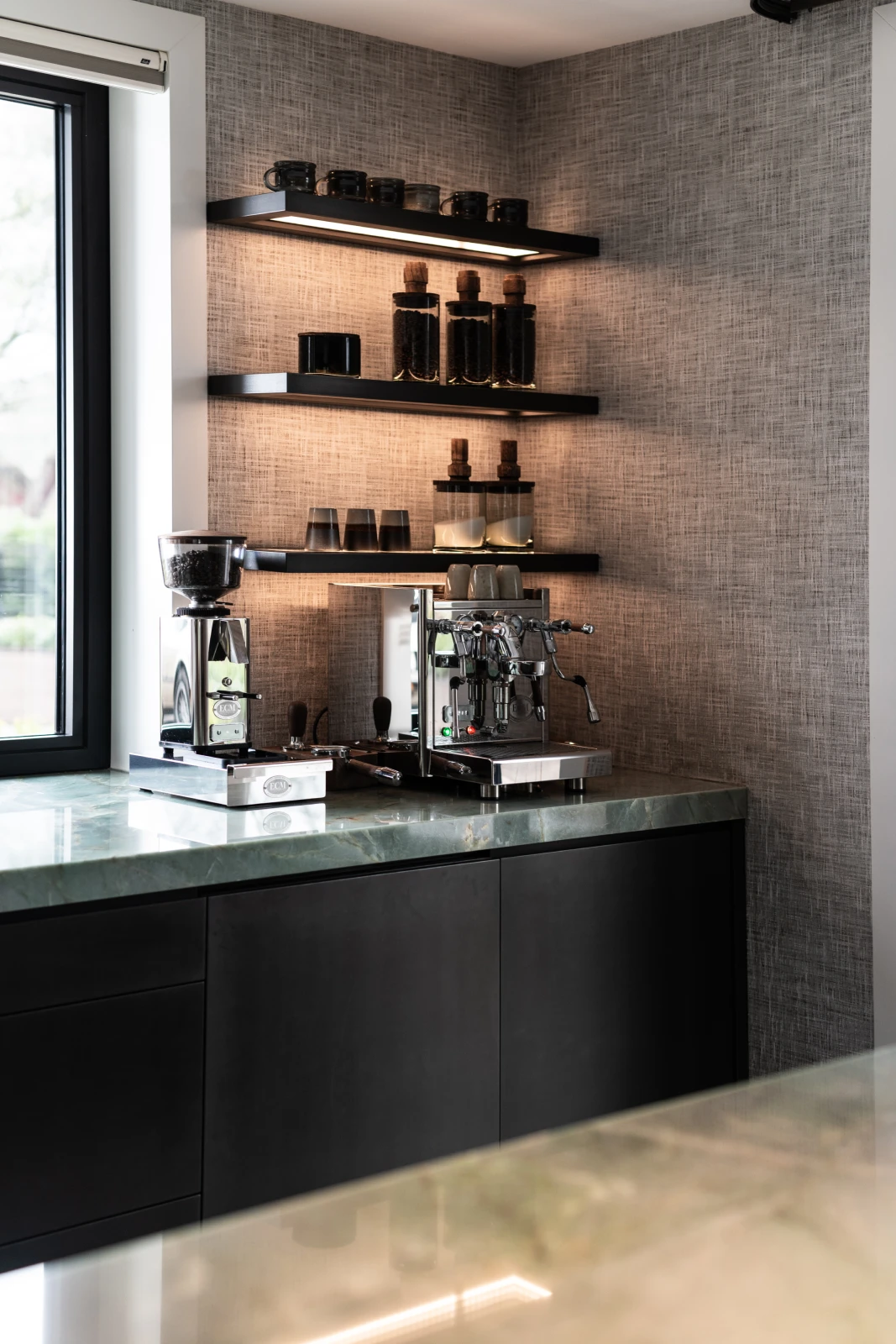 Een moderne koffiehoek met een koffiezetapparaat, molen en kopjes op een marmeren aanrecht onder zwevende planken met potten, mokken en glazen. Natuurlijk licht valt naar binnen door een raam aan de linkerkant.