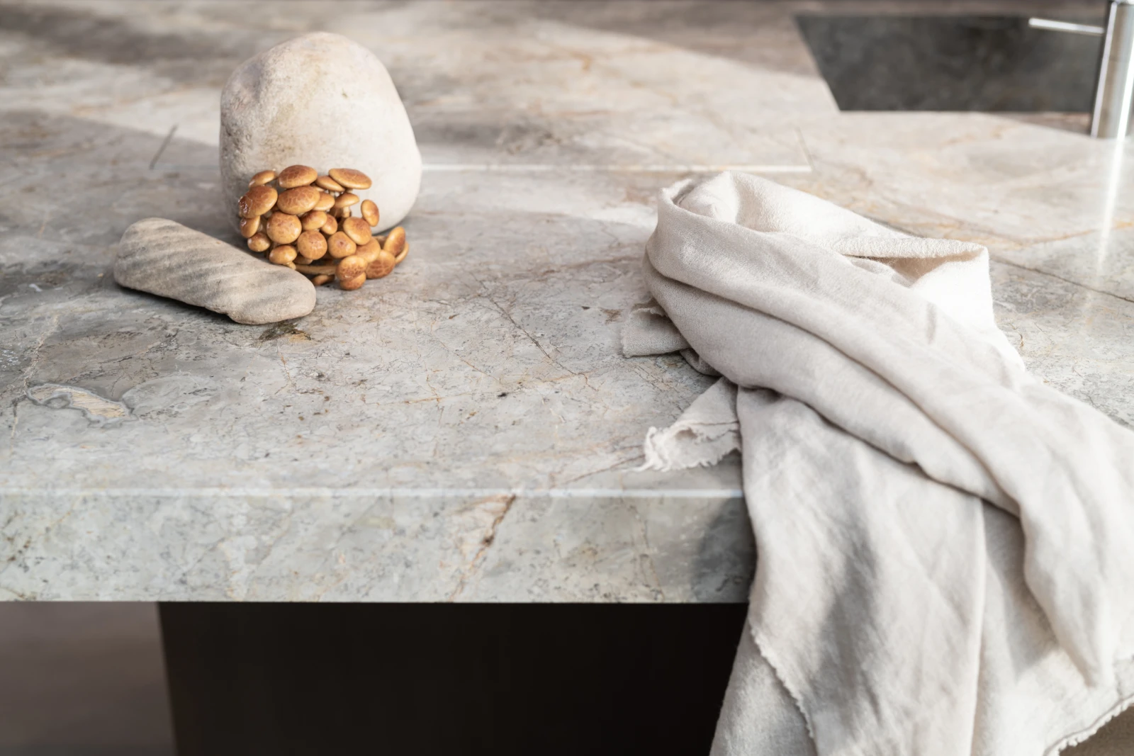 Een lichtgrijs marmeren keukenblad toont een beige steen, een geribbelde steen, geclusterde eikels en een lichte stof gedrapeerd langs de rand bij een ingebouwde wastafel.
