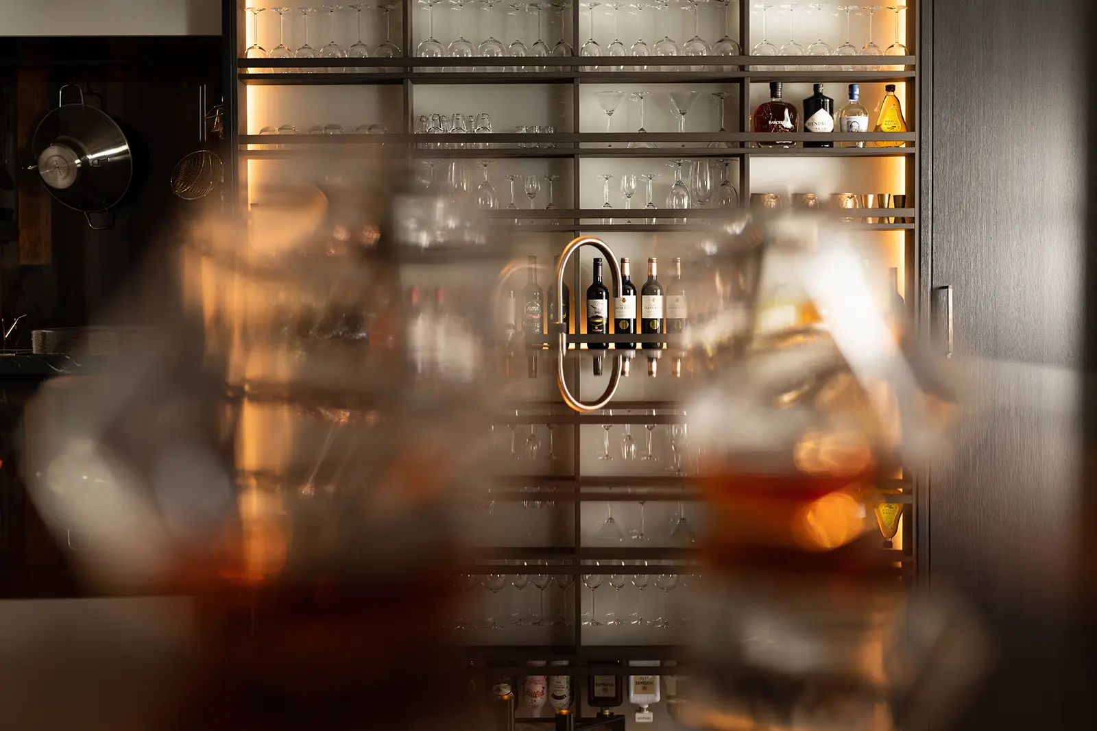 Een wazige voorgrond met glazen amberkleurige vloeistof, gericht op een moderne verlichte barplank met wijnflessen, divers glaswerk en flessen sterke drank.