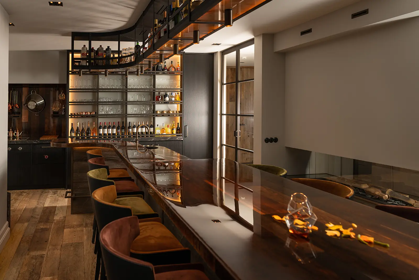 Een eiken, warm verlichte huisbar heeft een gepolijste houten toonbank en fluwelen barkrukken, met planken vol flessen en glazen. Een granieten keukenblad voegt verfijning toe aan de houten vloer en de strakke, minimalistische inrichting.