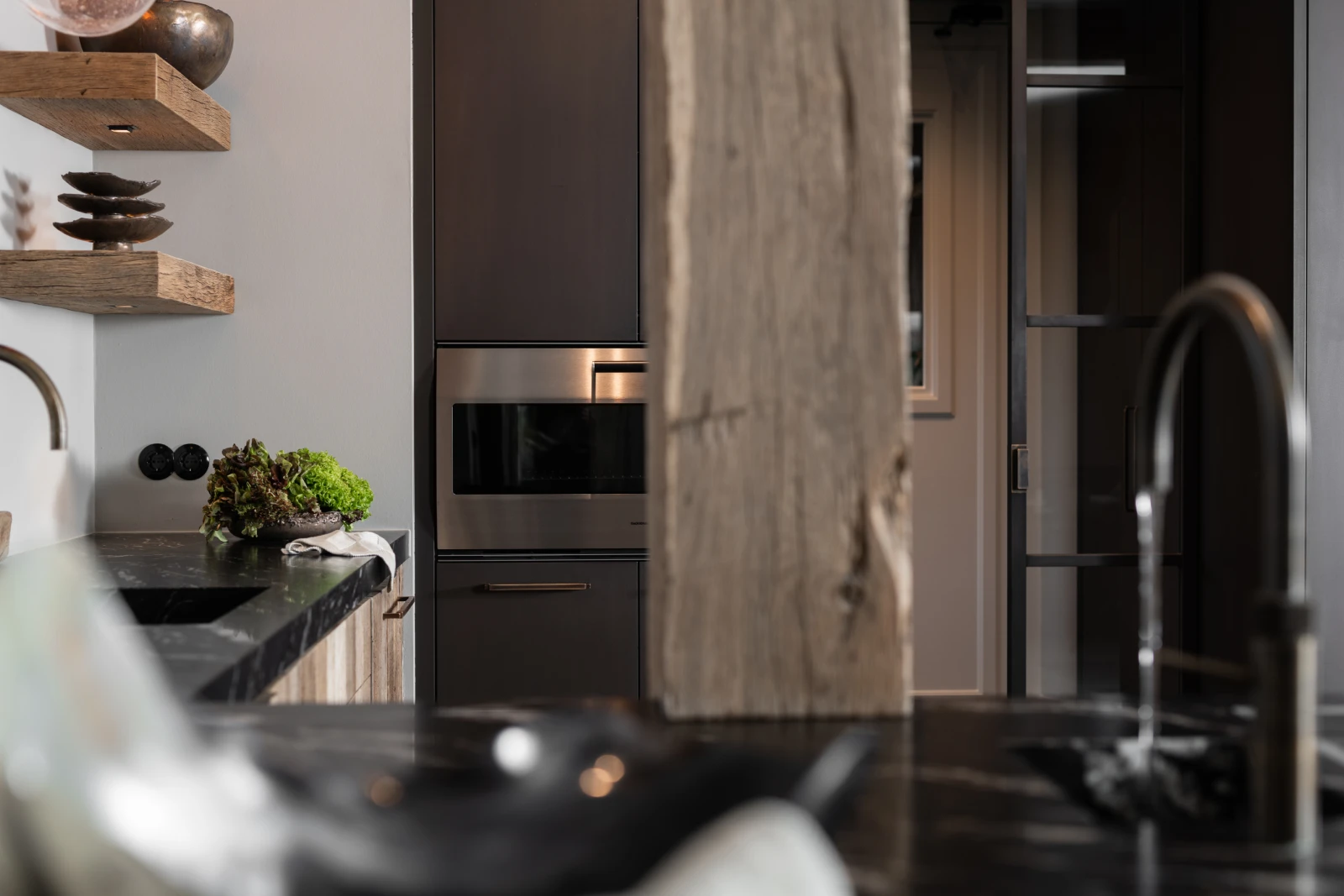 Een moderne oud eiken keuken met donkere kastjes, een ingebouwde magnetron, open houten planken met decor, een granieten keukenblad met een gootsteen en een rustieke houten balk in het midden.