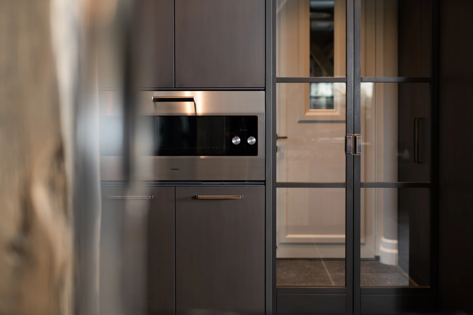 Een moderne keuken met donkere kastjes, een ingebouwde roestvrijstalen oven en glazen schuifdeuren die leiden naar een lichtgekleurde hal met een tegelvloer en een klein raam.