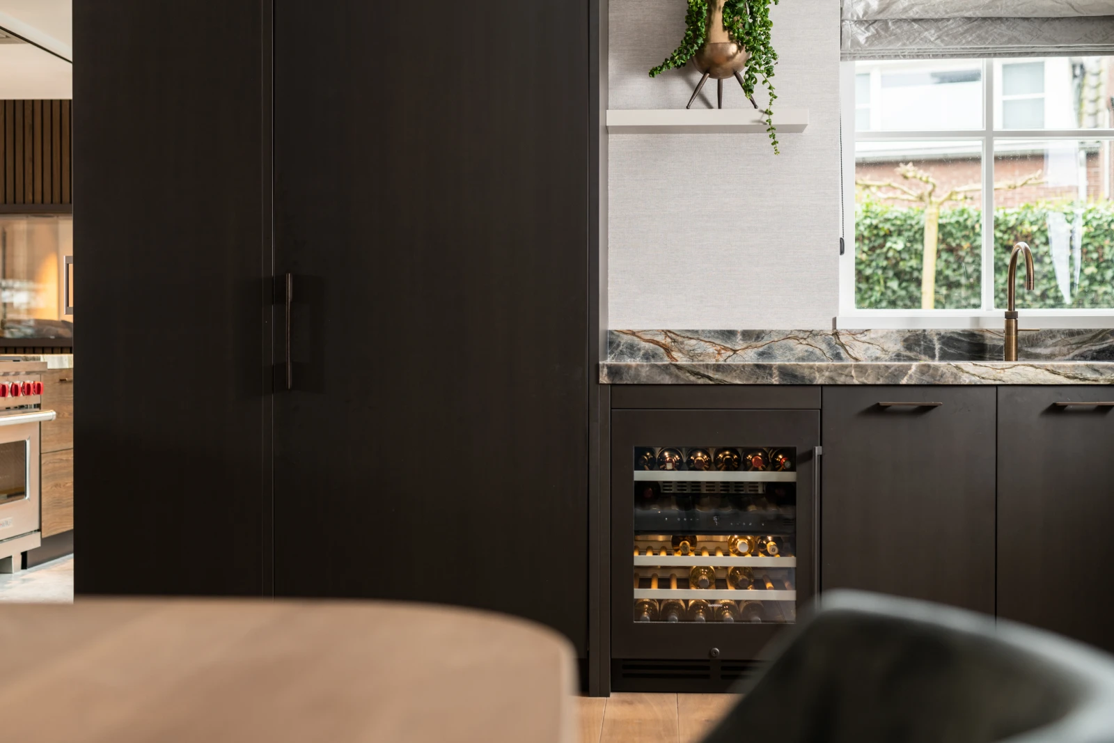 Keuken met donkere kasten, een kwartsiet aanrechtblad en een ingebouwde wijnkoelkast onder het aanrecht. Een raam boven de gootsteen laat natuurlijk licht binnen en er staat een plantje op een witte plank.