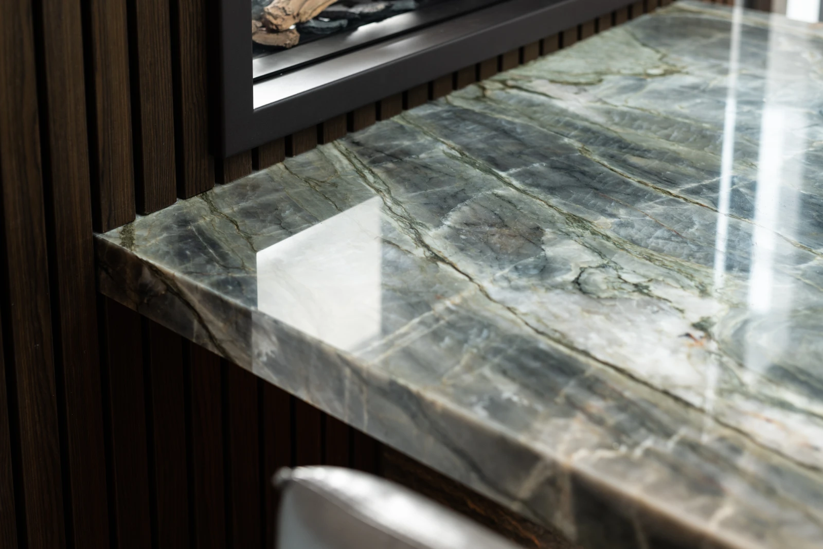 Een close-up van een werkblad in kwartsiet montagne blue naast een wand van donker verticaal houten panelen en een reflecterende metalen stoel. Licht wordt gereflecteerd op het gladde marmeren oppervlak.