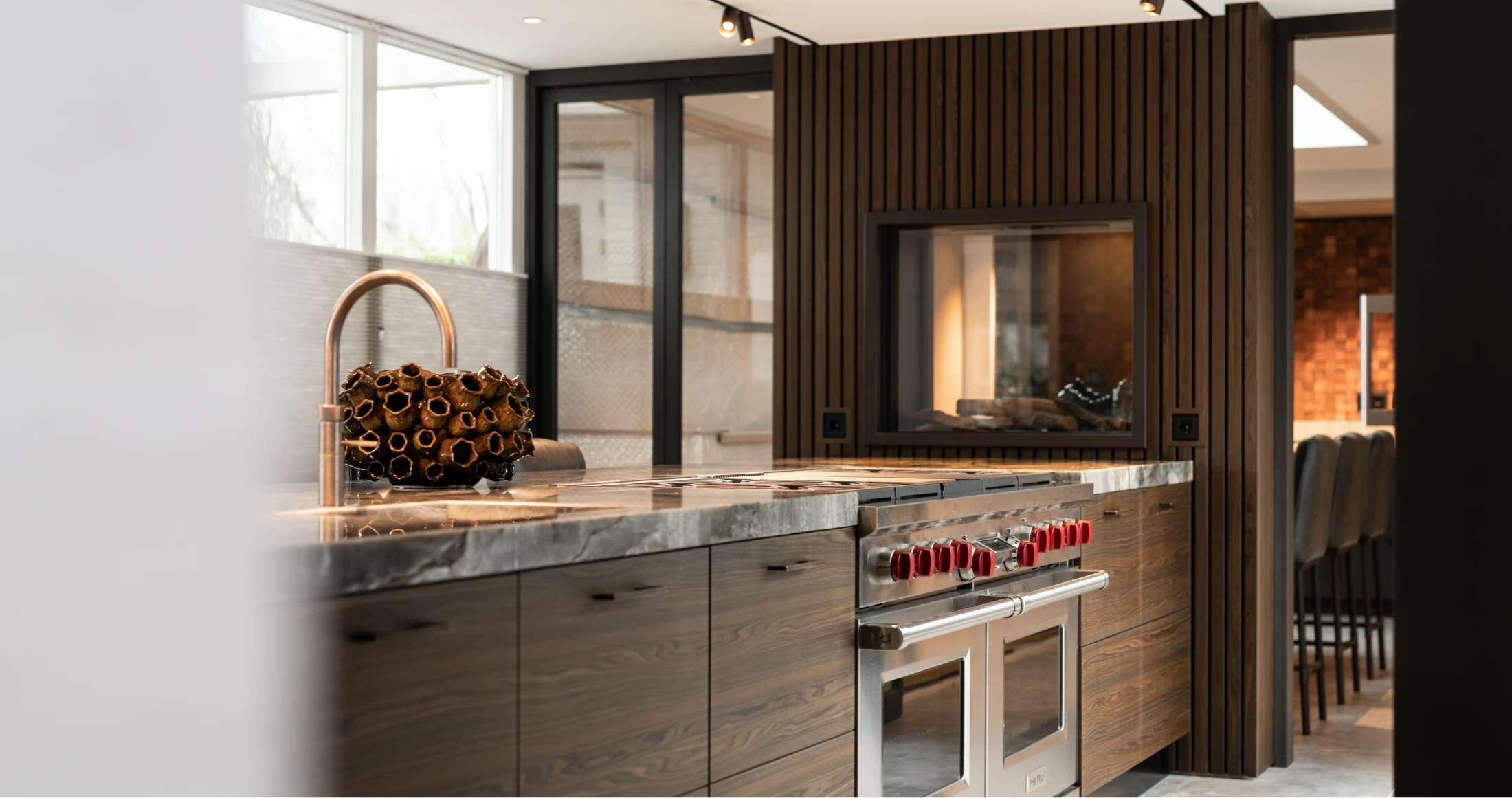 Moderne keuken met donkerhouten kasten, een marmeren aanrechtblad, Wolf apparatuur waaronder een roestvrijstalen fornuis met rode knoppen en een decoratieve mand. Grote ramen en verticale houten lambrisering geven de ruimte licht en warmte.