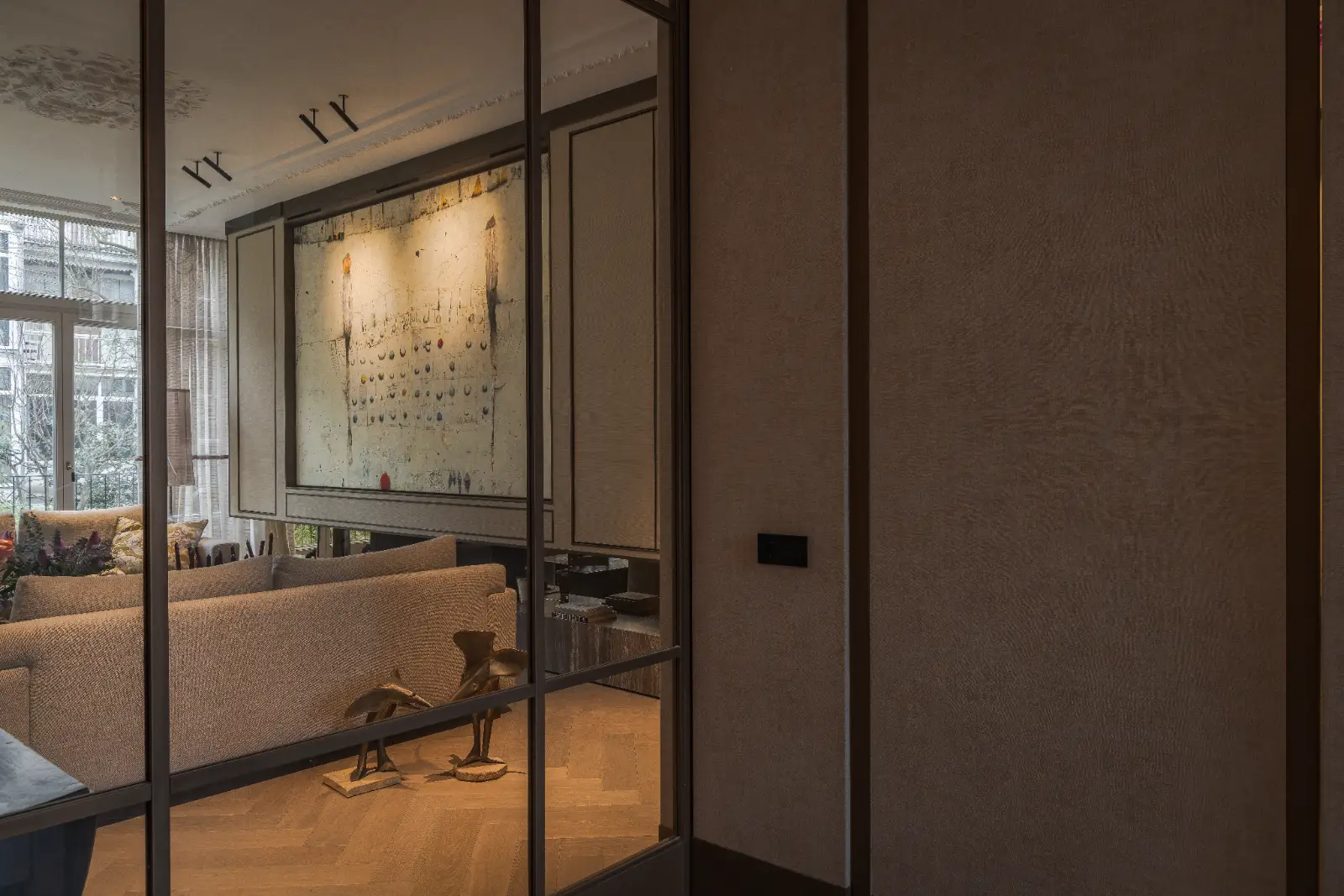 Een moderne woonkamer gezien door glazen deuren, met een beige sofa en abstracte muurkunst. Dit elegante herenhuis interieur wordt versterkt door zachte, warme verlichting, een kleine tafel met decor en grote ramen die natuurlijk licht binnenlaten.