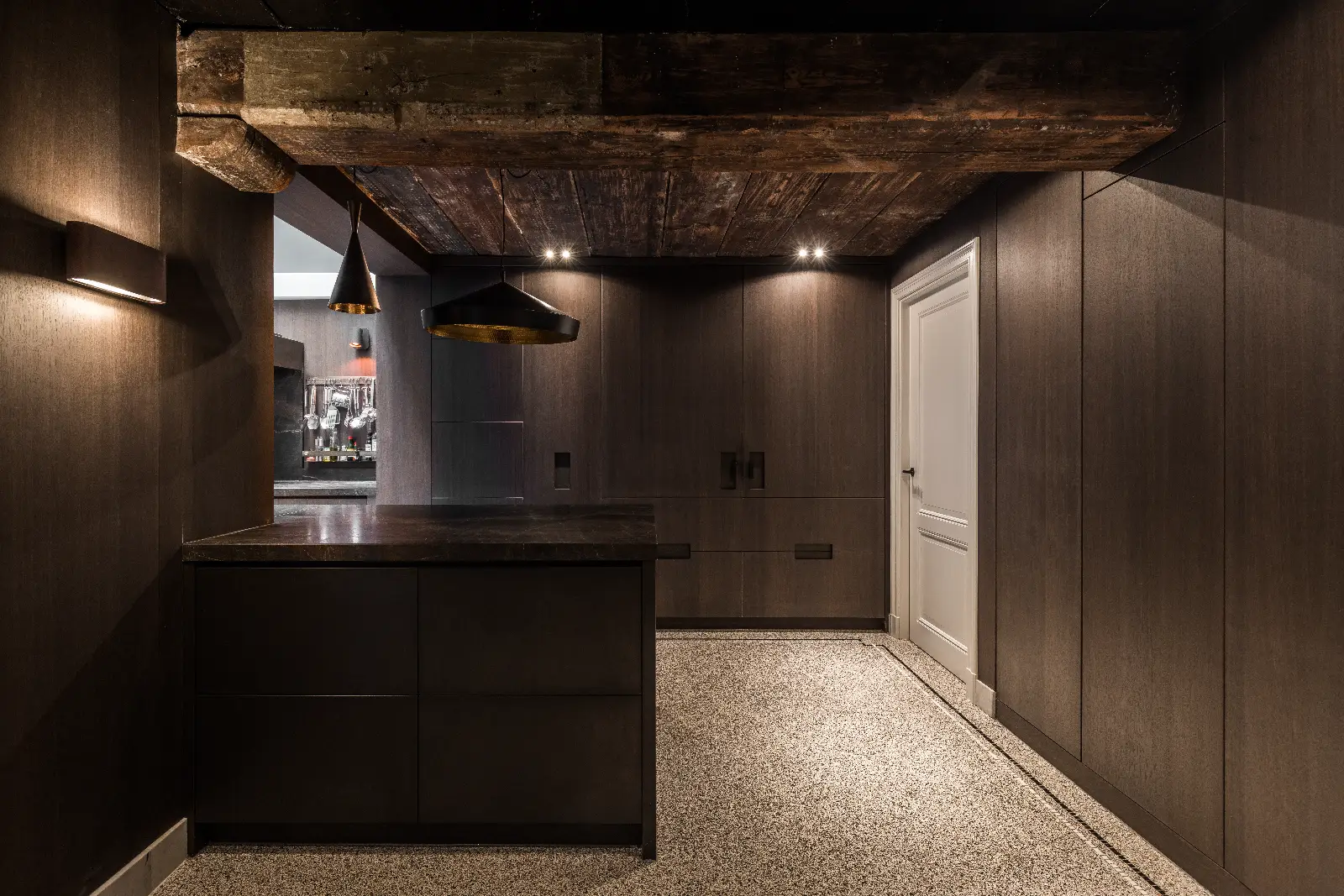 Minimalistische moderne keuken met donkerhouten kasten, bruine plafondbalken met structuur, zwart aanrechtblad en hanglampen. Dit verfijnde herenhuis interieur heeft een gesloten witte deur, een gespikkelde vloer en warme muurverlichting voor een gezellige touch.