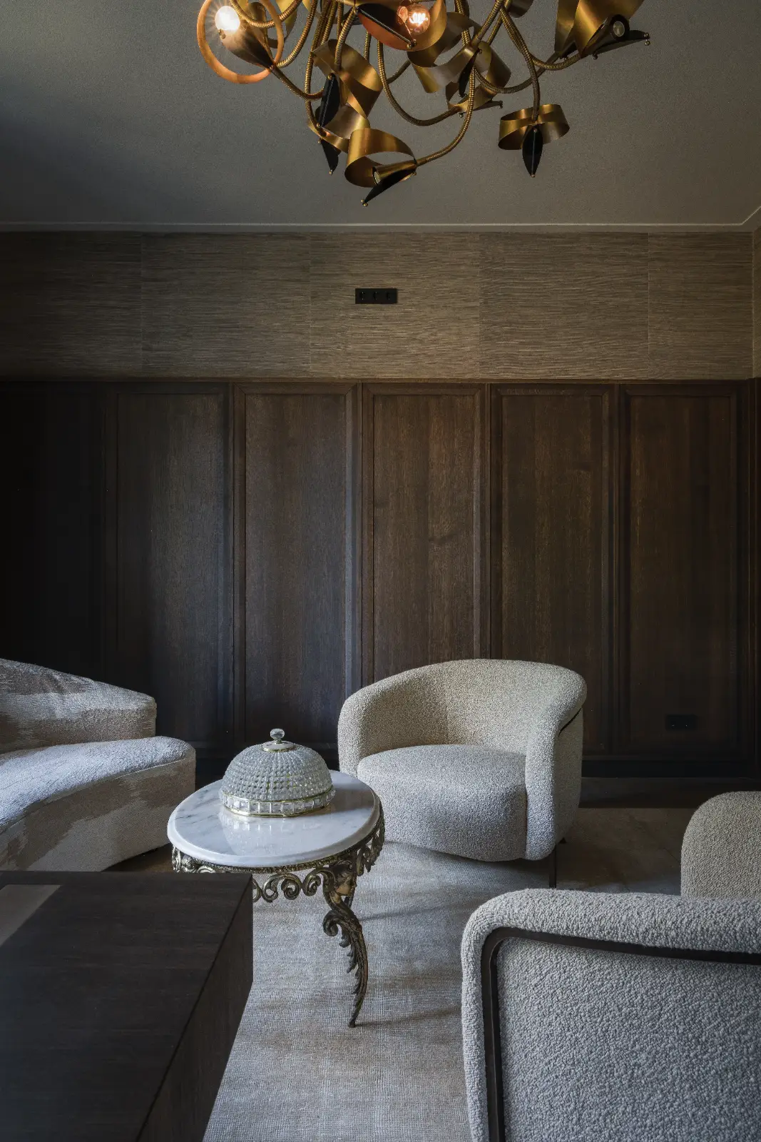 Een moderne woonkamer met muren van donker houten panelen, twee gebogen beige fauteuils, een wit marmeren salontafel met sierlijke gouden poten en een sculpturale gouden kroonluchter boven het hoofd - perfect voor de inrichting herenhuis.