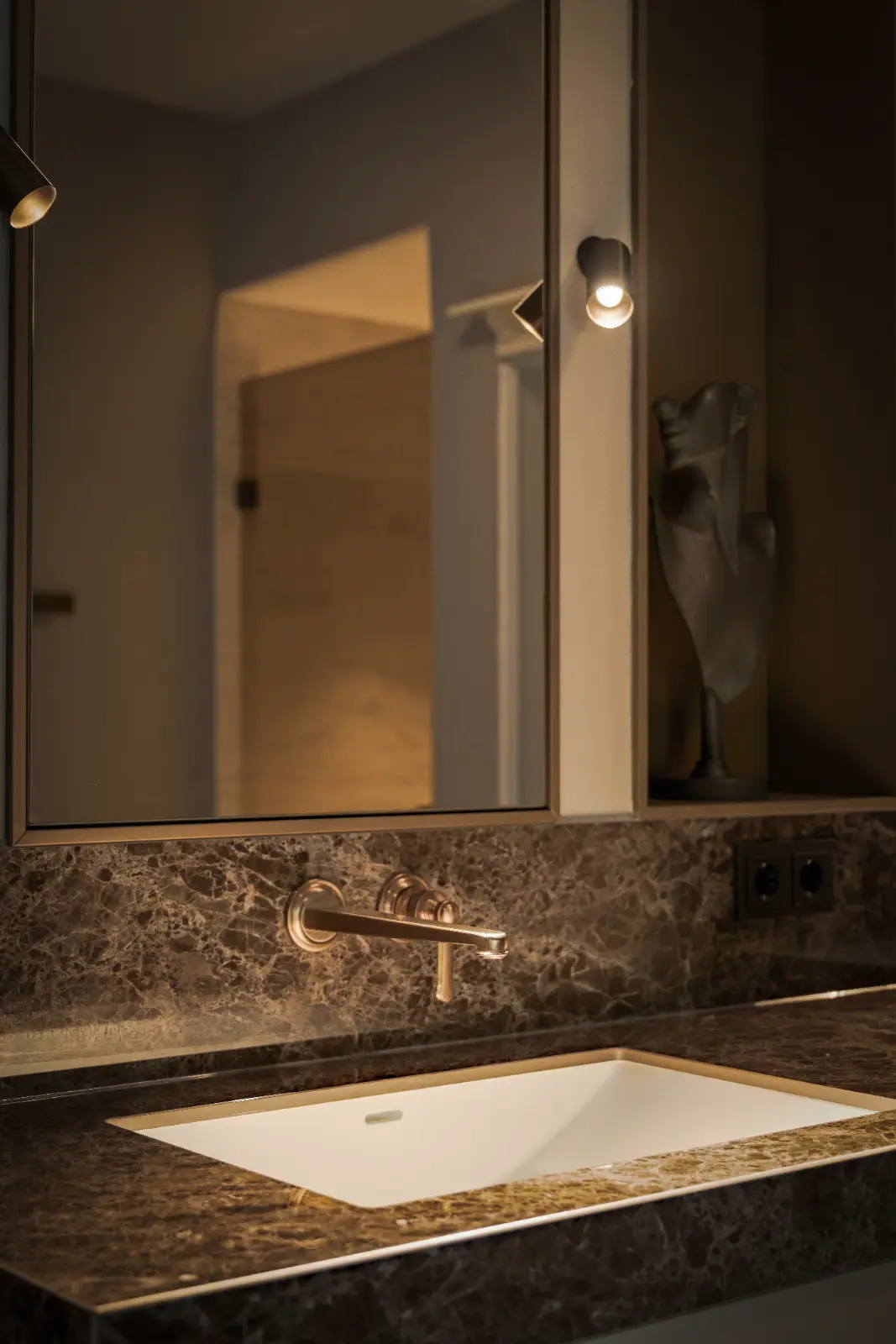 Een moderne badkamerwastafel met een marmeren aanrechtblad, een wandgemonteerde kraan en een grote spiegel wordt zacht verlicht, waardoor een serene sfeer ontstaat. Naast de spiegel rechts staat een decoratieve sculptuur, perfect voor herenhuis interieur.