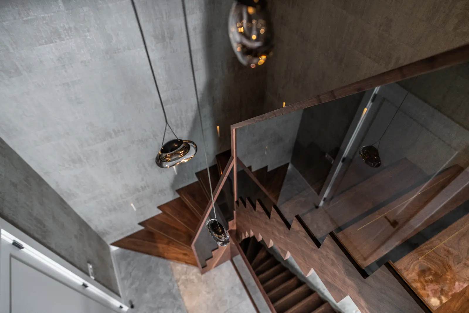 Een moderne houten trap met glazen leuningen is van bovenaf gefotografeerd. De grijze gestructureerde muren en hangende hanglampen voegen een strakke, eigentijdse toets toe aan dit minimalistische luxe villa-interieur.