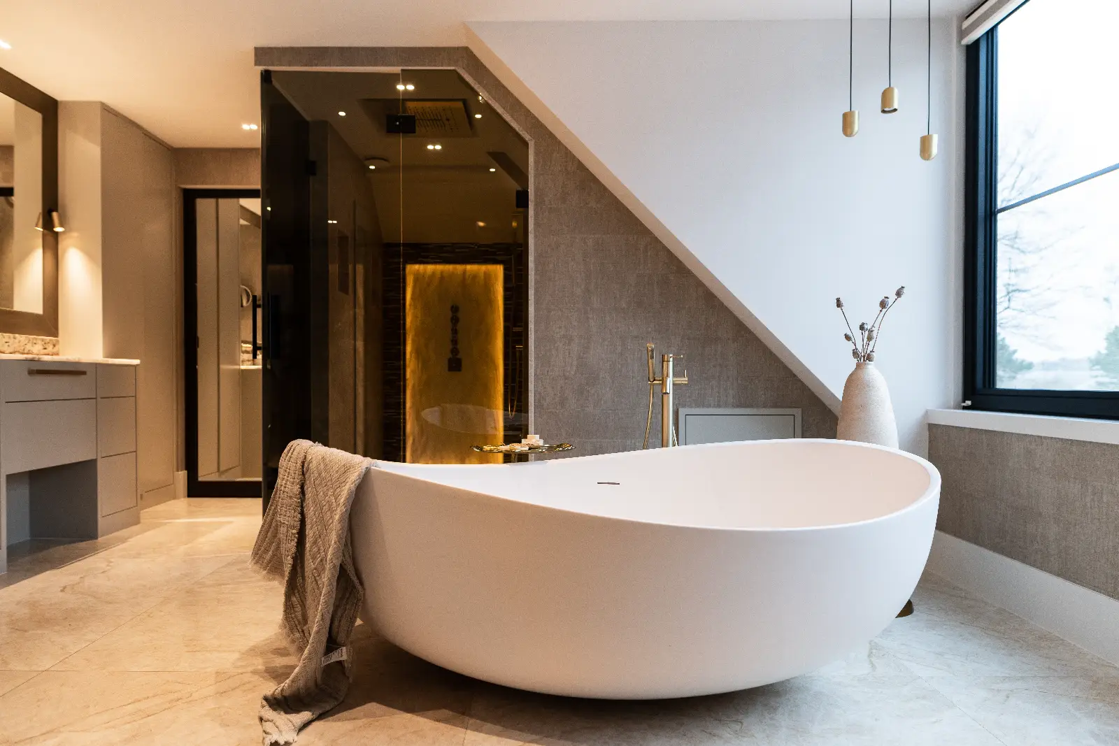 Moderne villa interieur badkamer met een vrijstaand wit ovaal bad met een gevouwen grijze handdoek, gouden armaturen, glazen douche, groot raam en neutraal getinte inrichting gevuld met natuurlijk licht.