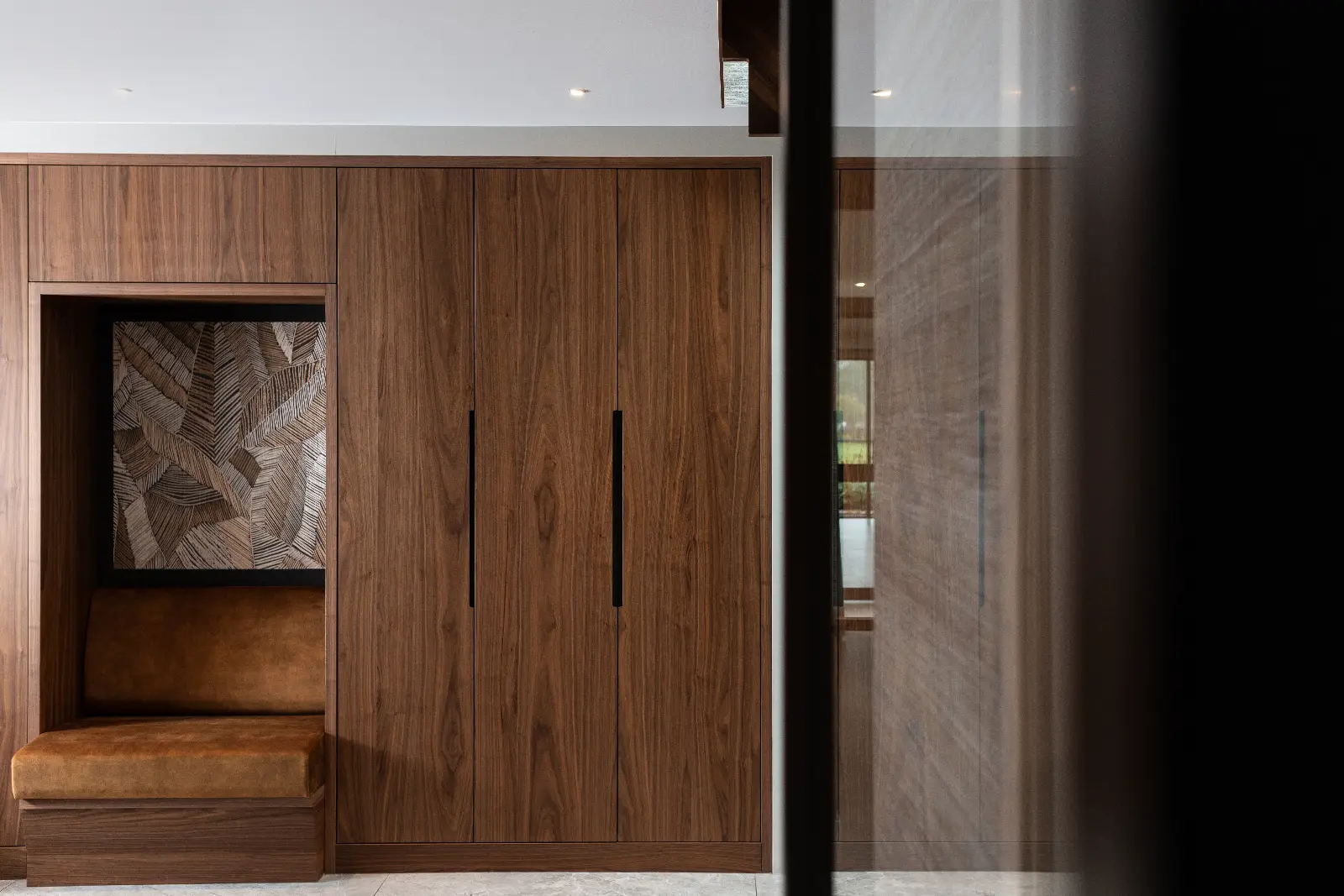 Een modern villa-interieur heeft ingebouwde houten kasten met verticale zwarte handgrepen, een bruine leren bank en een ingelijst decoratief bladmotief boven de bank. De ruimte straalt een strak, minimalistisch ontwerp uit.