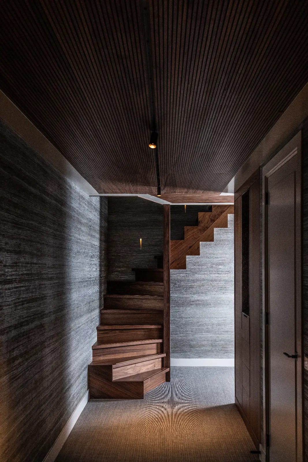 Een moderne, schemerige hal met donkere muren en plafond met textuur, die de villa interieurstijl laat zien - een unieke houten trap met geometrische treden leidt naar boven, terwijl rechts een gesloten witte deur zit.