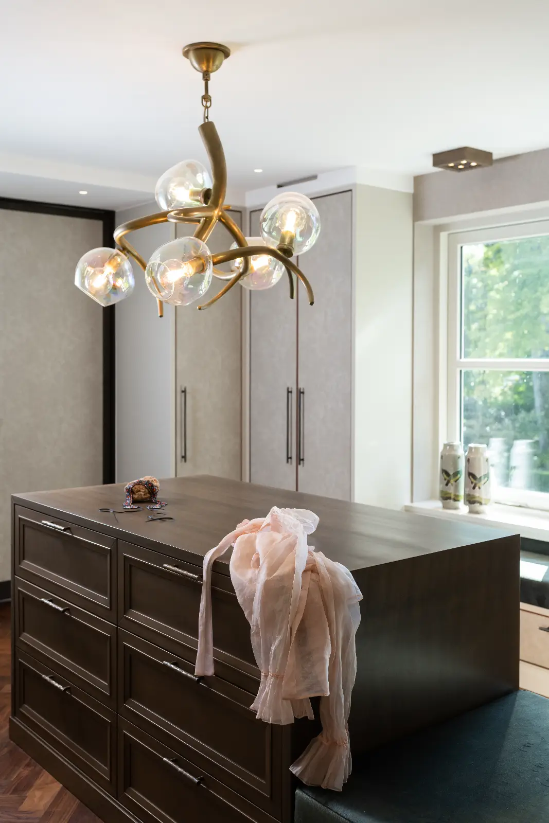 Een moderne inloopkast met een eiland van donker hout, een roze sjaal eroverheen gedrapeerd en een decoratieve lamp verbeeldt de elegante villa interieurstijl, terwijl het zonlicht door een raam met vazen op de vensterbank stroomt.