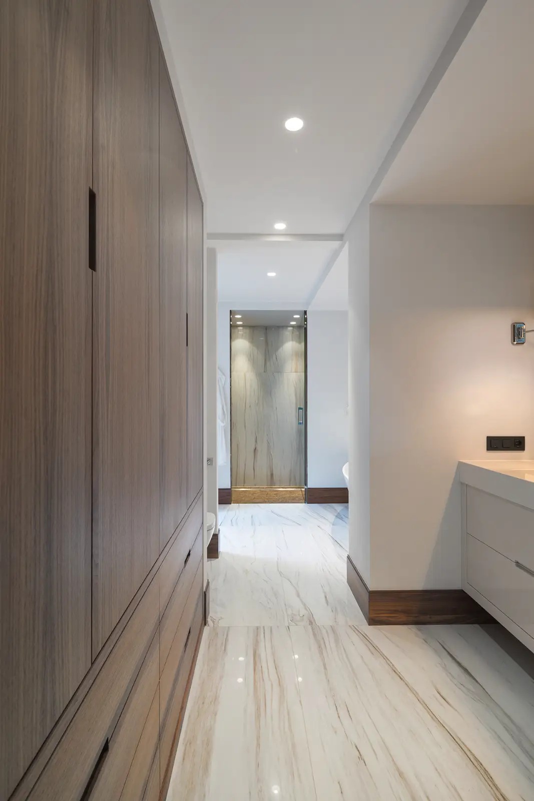 Moderne badkamer met marmeren vloeren, een grote houten inbouwkast aan de linkerkant, een wit badmeubel aan de rechterkant, inbouwverlichting en een glazen douchewand aan het einde van de gang - een luxe villa interieurstijl.