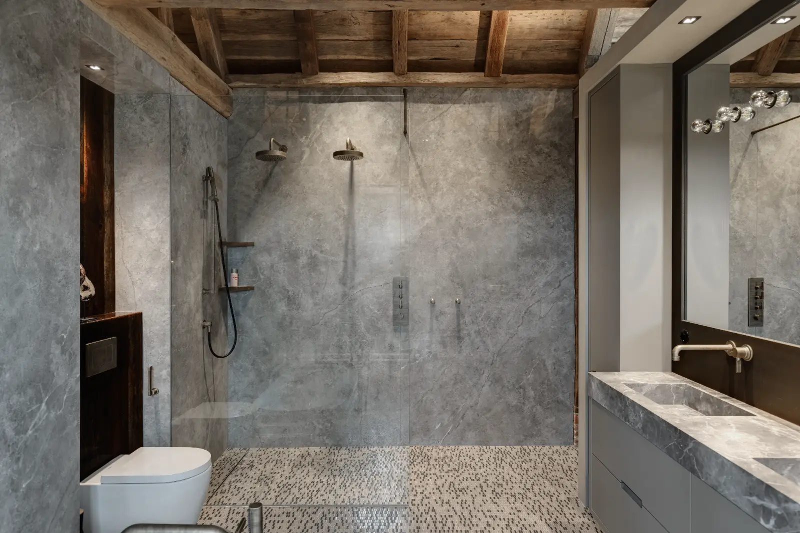 Moderne badkamer met grijs marmeren muren en vloer, open inloopdouche met dubbele douchekoppen, glazen scheidingswand, wandtoilet en grote spiegel boven een marmeren wastafel - perfect voor een ontwerp woonboerderij met zichtbare houten balken die rustieke charme toevoegen.