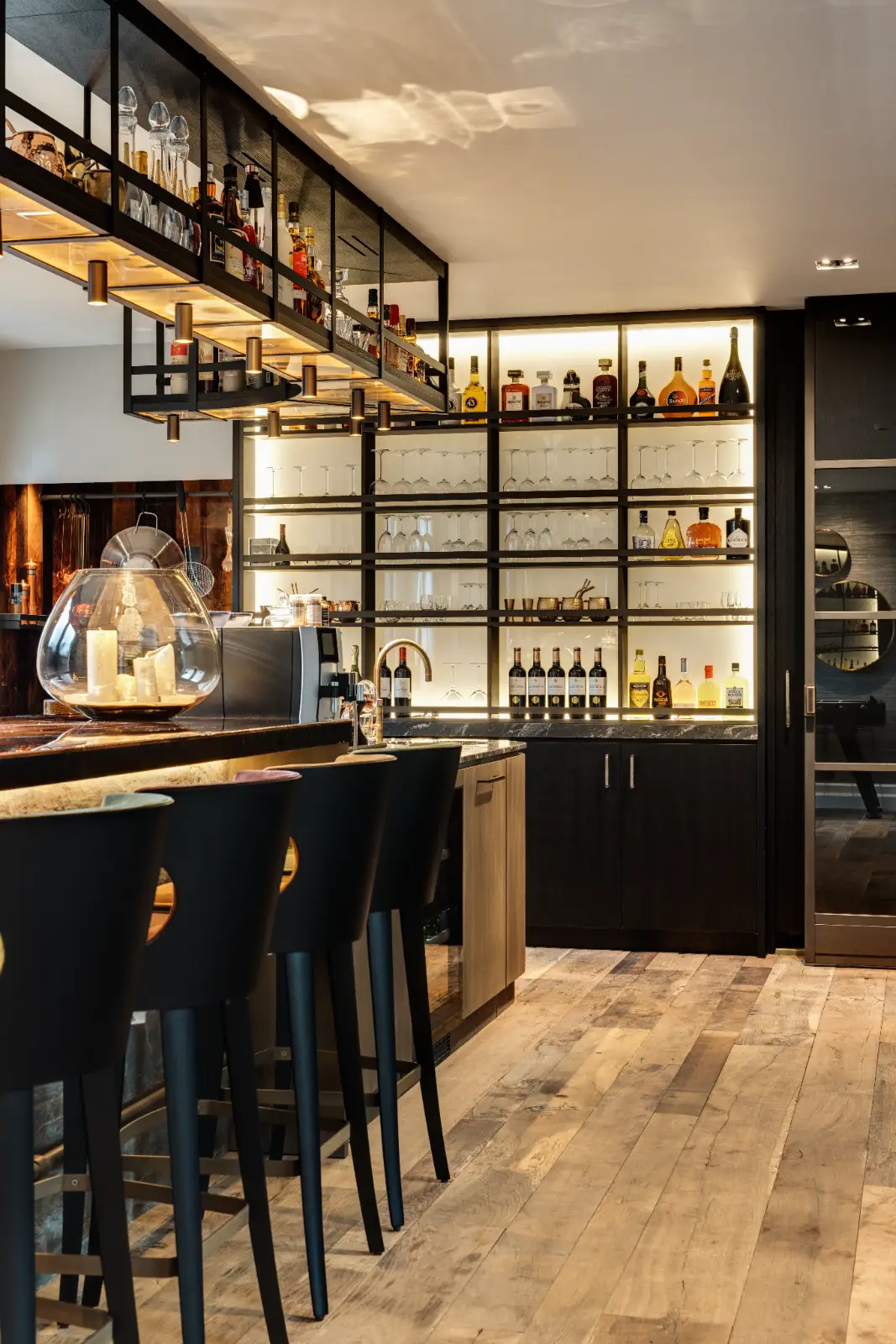 Een moderne huisbar met hoge zwarte stoelen, een marmeren aanrecht en planken met verschillende flessen drank en glaswerk, tegen warme verlichting en houten vloeren.