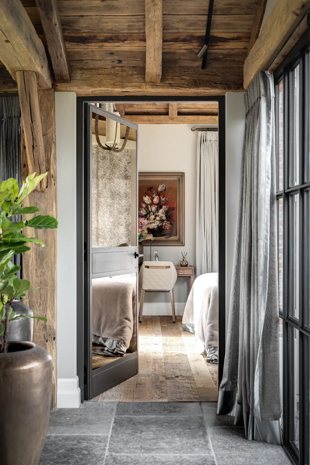 Een gezellige slaapkamer met rustieke houten balken, een zacht bed, neutraal beddengoed en een bureau onder een ingelijst bloemenschilderij geeft de charme weer van een ontwerp woonboerderij. Grijze gordijnen en een potplant voegen warmte toe terwijl het natuurlijke licht door de grote ramen binnenvalt.