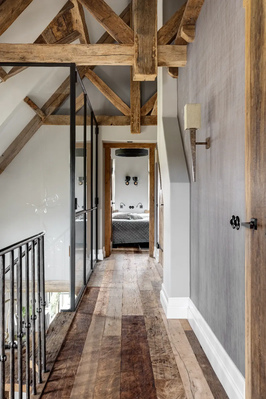 Een hal met rustieke houten balken en vloeren, grijze muren, deuren met glaspanelen en een zwarte metalen balustrade. Deze interieur woonboerderij heeft een badkamer met dubbele wastafels en spiegels aan het einde van de hal.