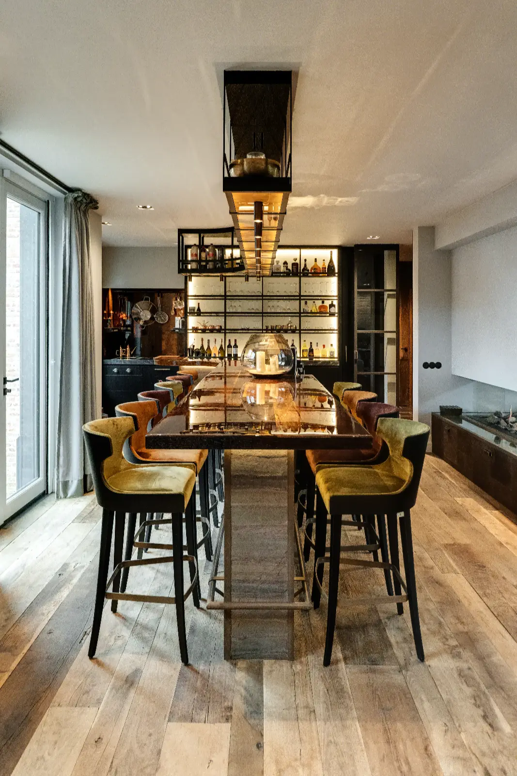 Een moderne eetkamer met een lange houten tafel, met fluweel beklede stoelen, planken met flessen en glaswerk, hangverlichting boven de tafel en een houten vloer. Deze stijlvolle interieur woonboerderij is gevuld met natuurlijk licht door glazen deuren aan de linkerkant.