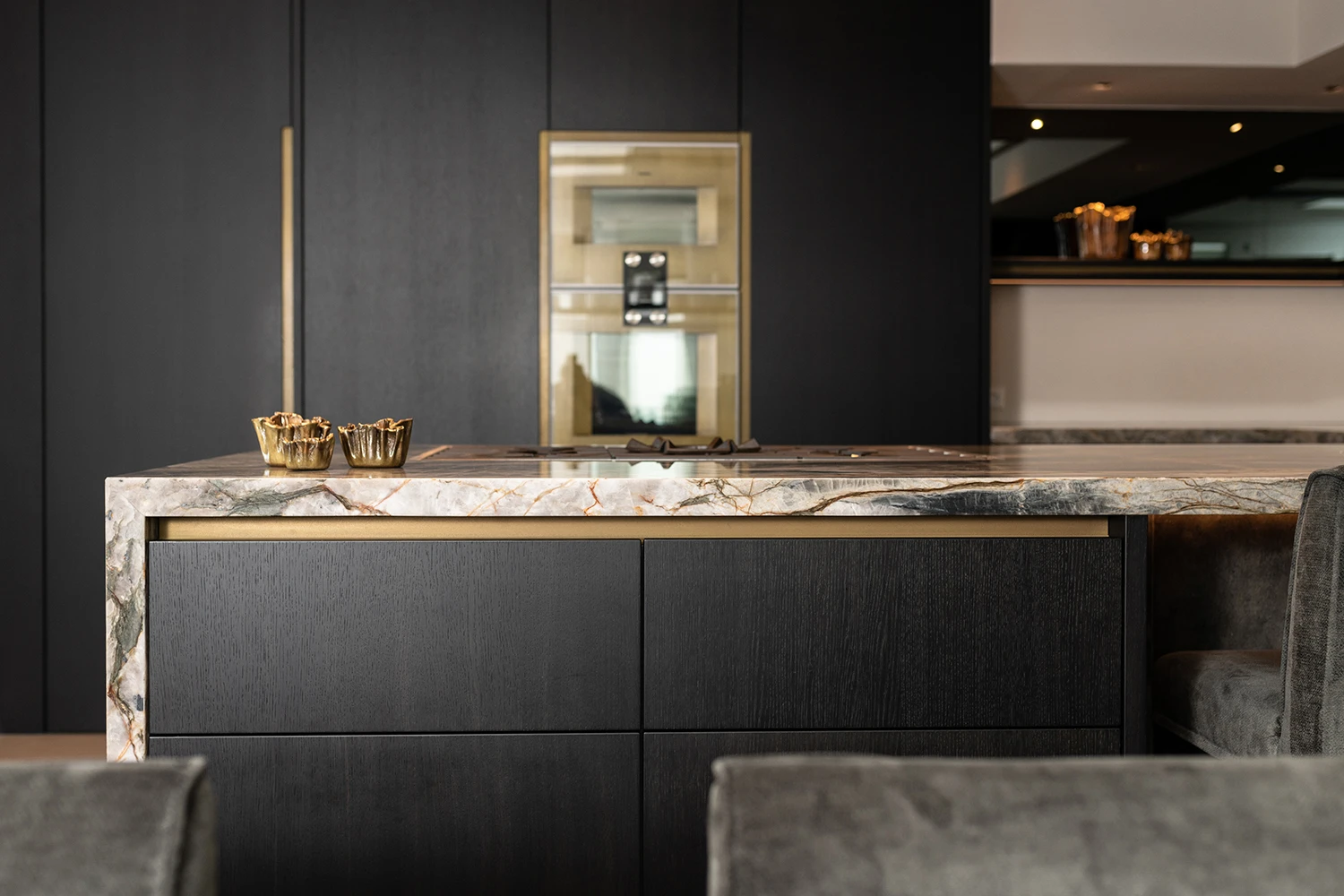 Moderne keuken van een keukenarchitect met een marmeren eiland, zwarte kasten, gouden accenten, ingebouwde oven en koffiezetapparaat. Decoratieve gouden schalen staan op het strakke, minimalistische eiland voor een geraffineerde touch.